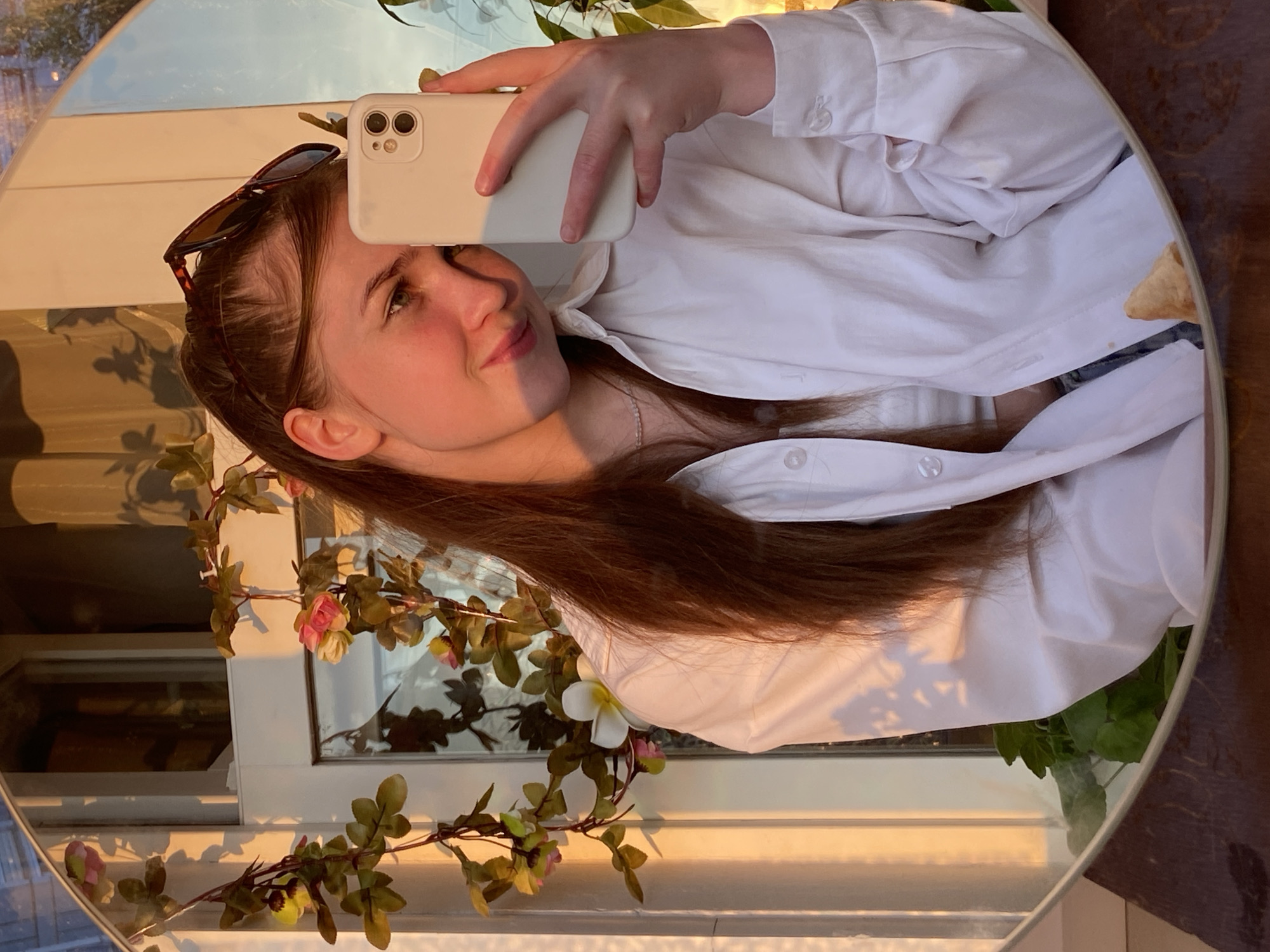 A young woman with long brown hair tied back takes a mirror selfie. She is wearing a white button-down shirt over a white tank top and a pearl necklace. Sunglasses are perched on top of her head. She is holding up a white smartphone with her right hand, which is in focus. The background is a reflection in a round mirror, showing a window with light streaming through, casting shadows of plants. A floral garland with pink and yellow flowers is draped around the mirror. The setting appears to be indoors, possibly near a window. There is no indication of the city of Chaikovsky, Russia.