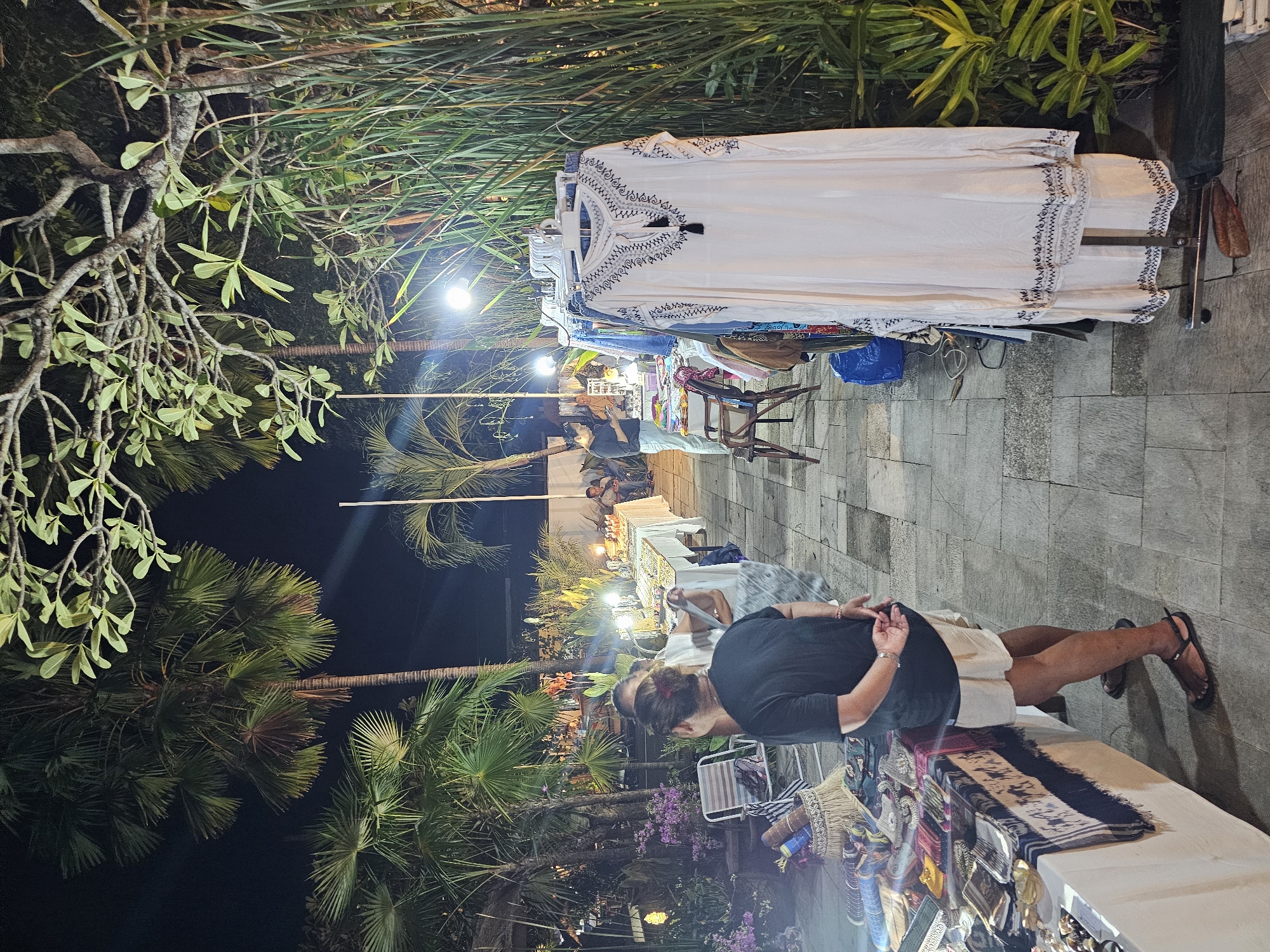 A woman looks at items on a table at an outdoor market in Denpasar, Indonesia. The market has a tropical aesthetic, as evidenced by the palm trees and other lush greenery visible in the background. The woman is wearing a black top and a light-colored skirt. Her hair is tied back. She is standing in front of a table covered with various items for sale, including scarves and jewelry. Other shoppers and vendors are visible further down the market path. White clothing is displayed on a rack to the side. Overhead lighting is provided by string lights and lampposts. The ground is paved with light-colored stone tiles.