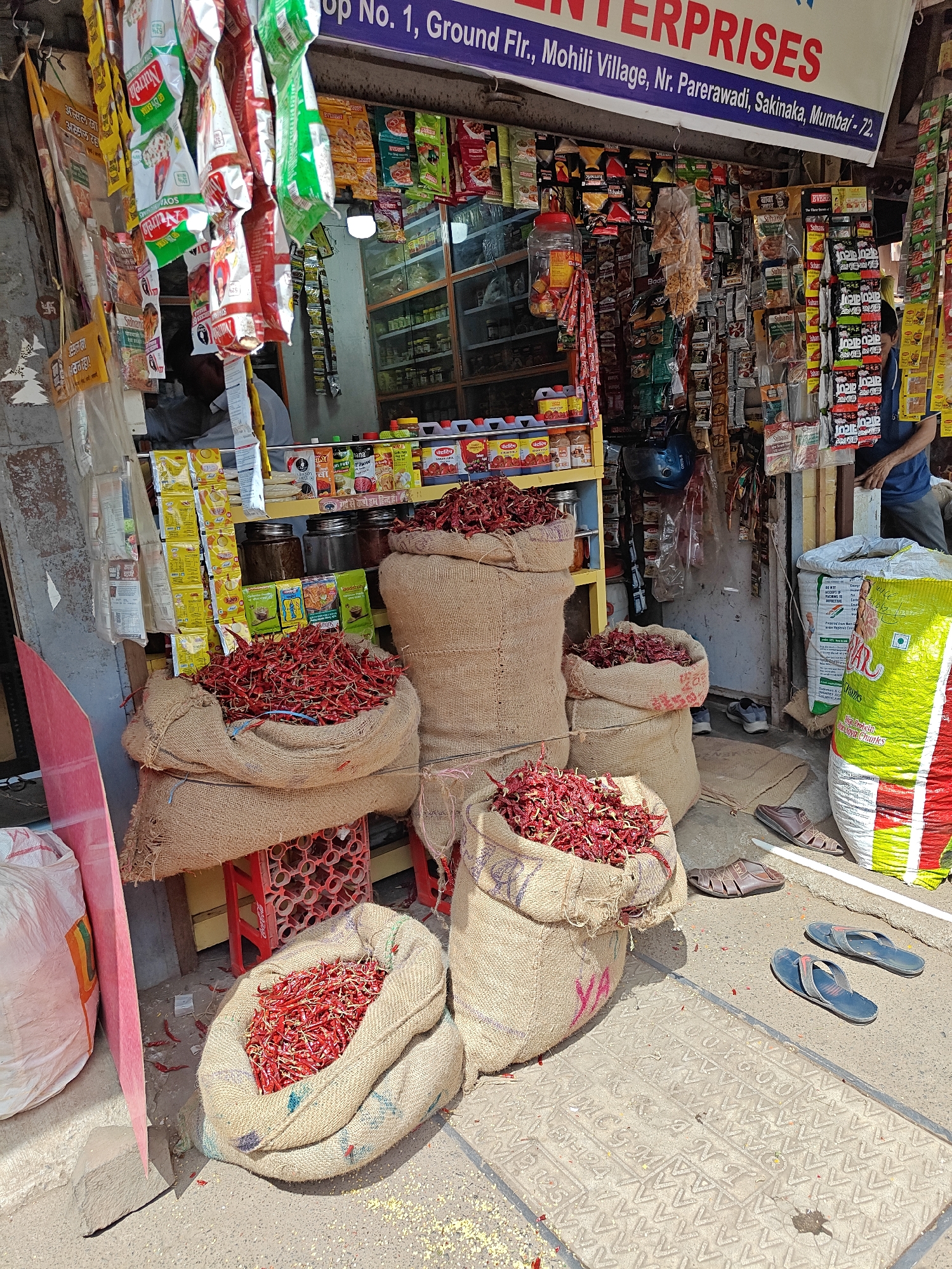 Here is a detailed description of the image:
**Scene:** An outdoor storefront, likely a small grocery or spice shop, filled with sacks of dried red chilies.
**Location:** The shop's banner indicates it's located at "Shop No. 1, Ground Flr., Mohili Village, Nr. Parerawadi, Sakinaka, Mumbai - 72."
**Subjects and Objects:**
* Five large sacks of dried red chilies in the foreground, displayed for sale, are piled up on the ground and elevated on small stools or crates.
* Behind the chilies is the shop's entrance. The shop is stocked with various food products, packaged snacks, spices, and everyday items, visible on shelves and hanging from the ceiling.
* To the side, is a yellow and green bag and a pink divider.
**Actions and Interactions:**
* A shopkeeper or assistant is visible standing inside the shop.
* Sandals and shoes are scattered around the bags of chilies, suggesting customers or passersby.
**Environment:**
* The area appears to be a bustling street or market area.
* The storefront is simple, with a basic awning and signage.
* There is some debris and scattered spices around the base of the chili sacks.