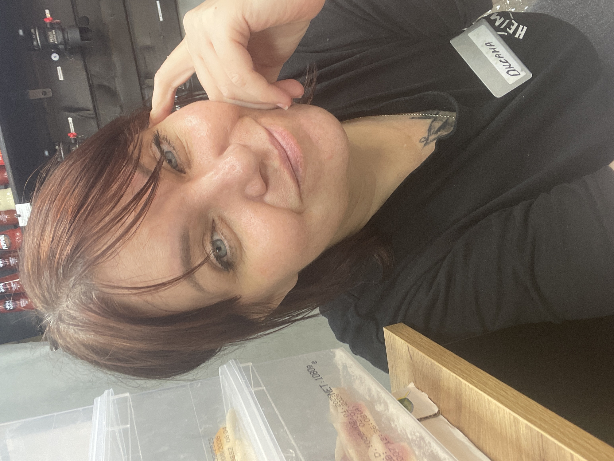A woman with auburn hair and blue eyes is taking a selfie in what appears to be a storage area or stockroom. She is wearing a black t-shirt with a name tag that reads "DICKANA" attached to it. She is resting her right hand on her cheek and smiling at the camera. In the background, there are clear plastic storage containers stacked on shelves. Some of the containers have labels with text, including dates and weights, such as "02/2025", "08/2026", "NET: 1080g". On a shelf behind her, there are bottles of what appear to be condiments or sauces. The environment suggests a retail or food service setting. The location of Rusanovo, Russia, is not visually indicated in the image itself, but is provided as context.