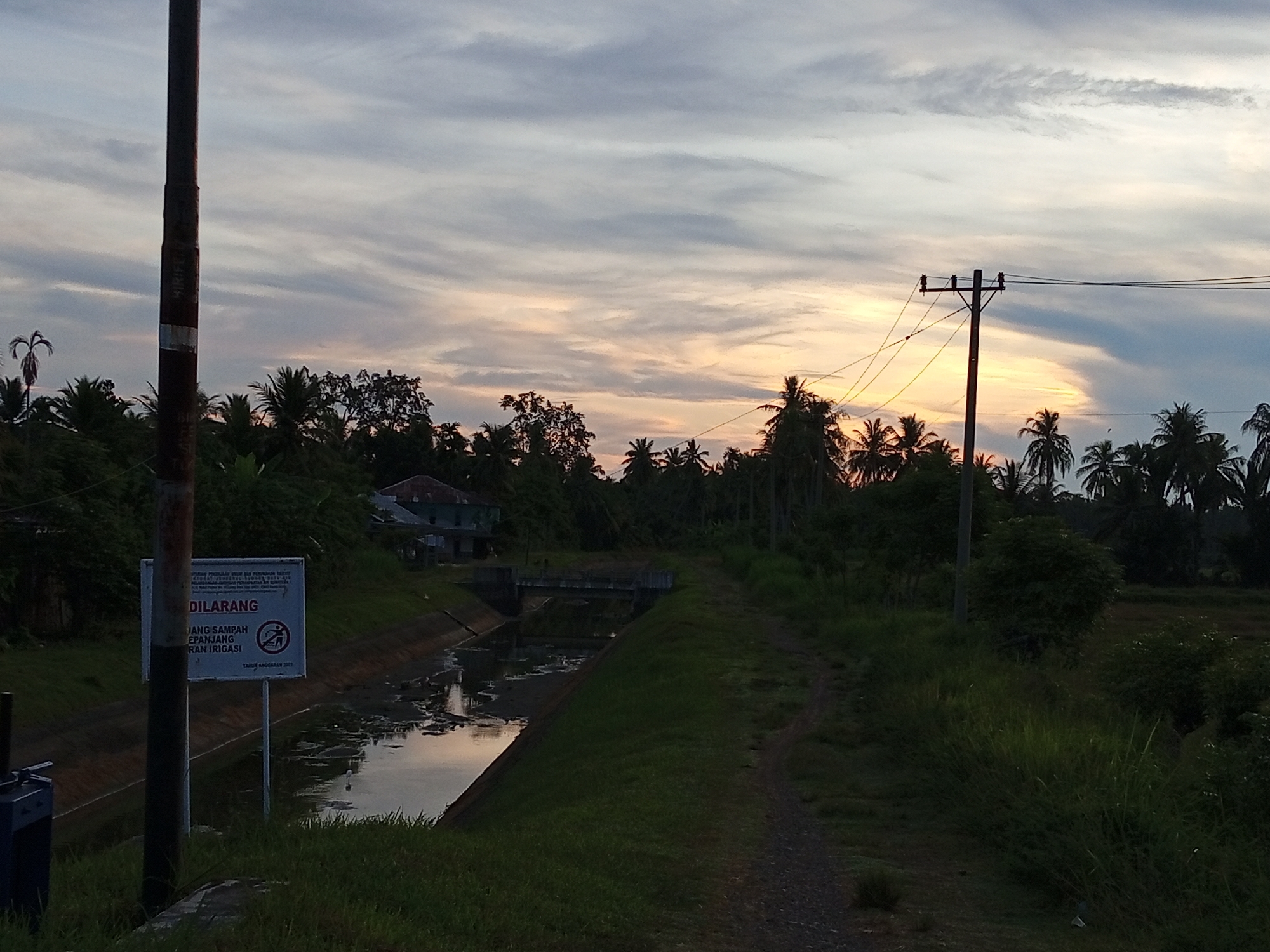 The image depicts a rural landscape at dusk in Cotjrat, Indonesia. A canal runs diagonally across the scene, with a path bordering its right side. Lush green vegetation, including palm trees, dominates the surroundings. In the foreground, a dark, weathered pole stands to the left, bearing a sign with Indonesian text and symbols. The sign appears to be a warning or prohibition, likely related to littering or waste disposal, as indicated by the word "DILARANG" (forbidden) and a pictogram of a person discarding trash. Beyond the canal, a small building with a red roof is visible amongst the trees, and further in the distance, a power pole with wires stretches across the frame against a cloudy sky. The sky displays a gradient of light, with hues of orange and yellow near the horizon, suggesting the setting sun. The overall scene is peaceful and natural, characterized by the interplay of water, vegetation, and the twilight sky.