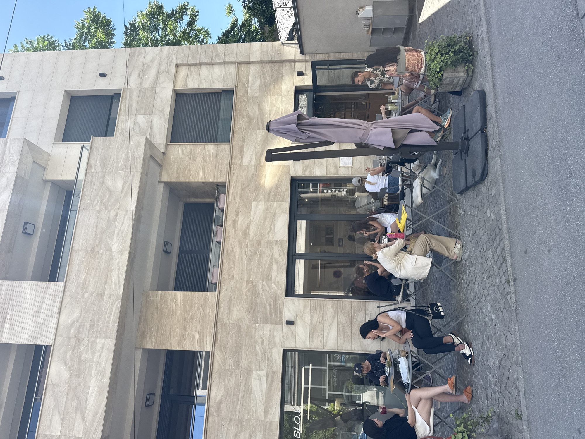 The image shows an outdoor café scene in Vracar Urban Municipality, Serbia. Several people are seated at tables outside a modern building with a light-colored, patterned facade. The building has large windows and closed blinds on the upper floors. People are eating, drinking, and conversing. A large umbrella provides shade for some of the tables. The street is paved with cobblestones. Plants in pots are placed near the café seating area.