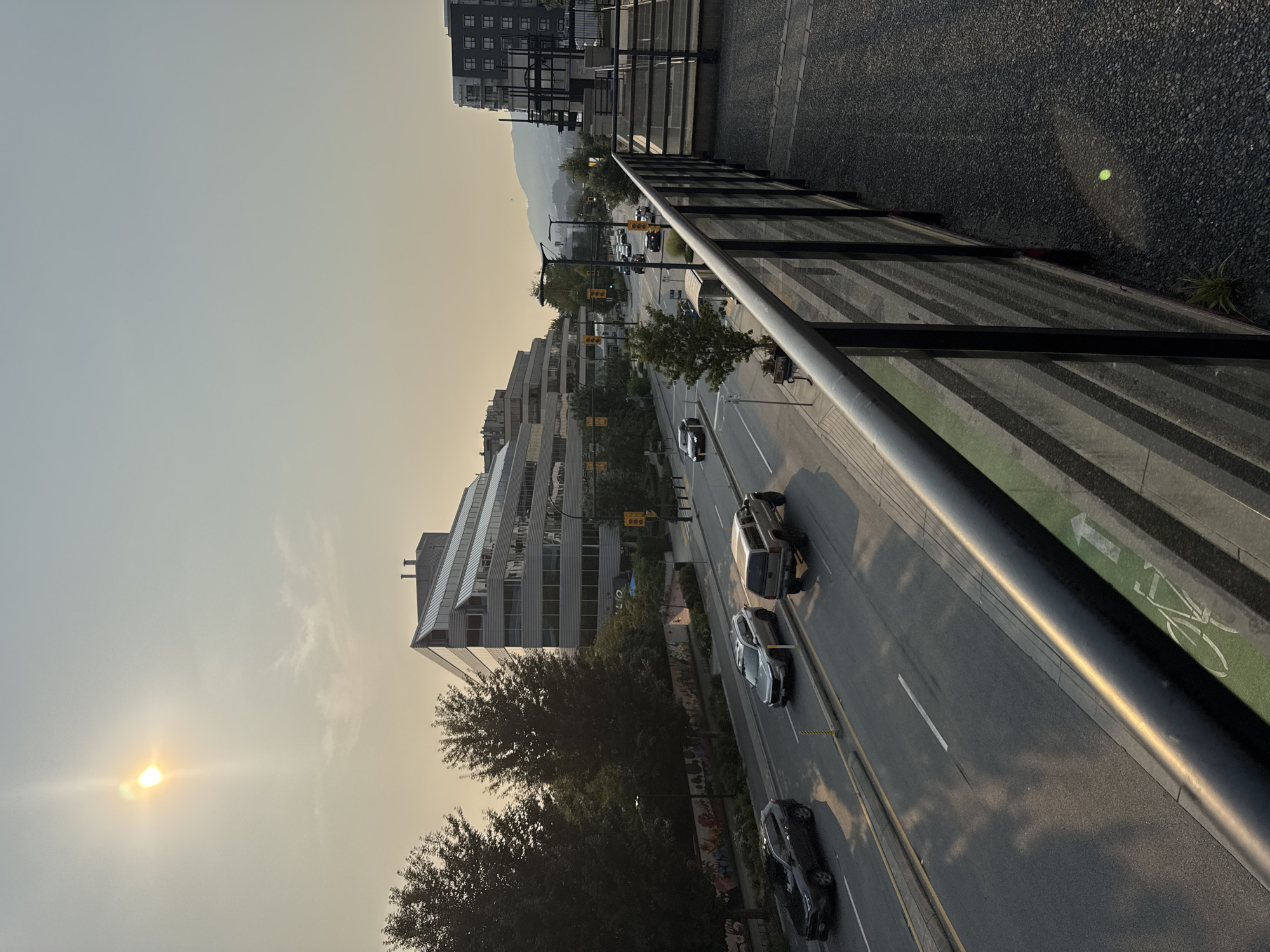 This is a daytime, eye-level, wide shot of a street scene in North Vancouver, Canada, likely during late summer or early fall due to the hazy sky and the warm, angled sunlight. The image is captured from an elevated perspective, possibly a balcony or elevated walkway, looking down onto a multi-lane road.
In the foreground, a dark metal railing with glass panels runs diagonally across the frame, casting long shadows. The ground beneath the railing is a textured, dark grey gravel.
The street itself is busy with traffic. Several vehicles are visible, including a black SUV, a silver SUV, and a pickup truck. The road has distinct lanes marked by white lines, and there is a green bike lane on the right side of the road, indicated by a bicycle symbol. Traffic lights are visible at an intersection further down the street.
On the left side of the road, a large, modern building with a distinctive stepped architectural design and a glass facade dominates the mid-ground. Trees with dense green foliage line the street in front of this building. A mural or artwork is visible on a wall behind the trees.
In the background, a hazy sky obscures distant mountains. The sun is visible high in the sky, partially obscured by haze, casting a bright glare and creating lens flare. Another, taller building with a dark facade and visible external scaffolding is seen on the right side of the frame, suggesting ongoing construction or development.
The overall impression is one of urban activity under a hazy, possibly smoke-filled, sky.