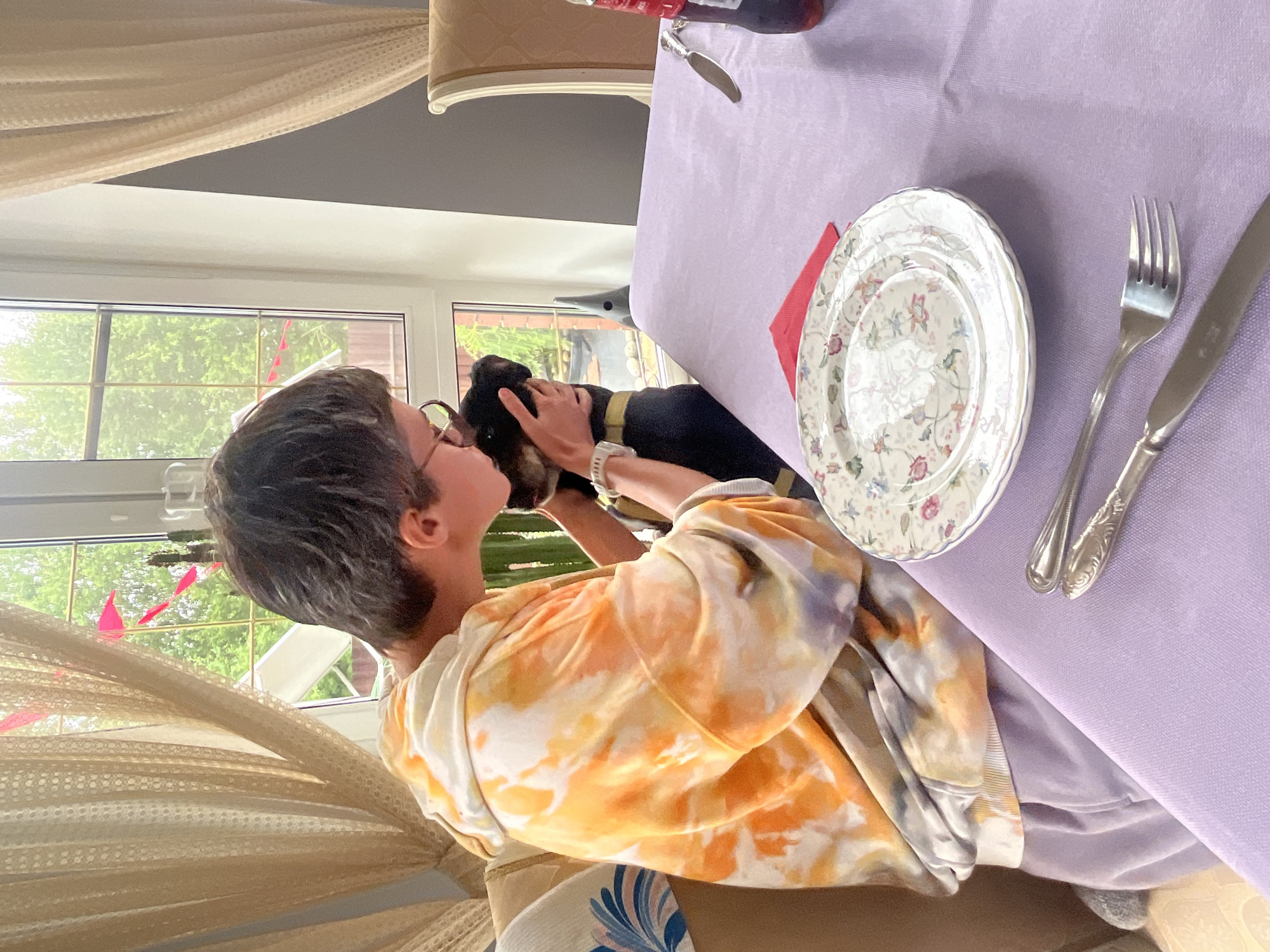 A person wearing a tie-dye hoodie and purple sweatpants is seen from the side, embracing a black and tan dog. They are seated at a table covered with a lavender tablecloth. On the table, there are two plates, one stacked on top of the other, both with a floral pattern. A red napkin is placed to the side of the plates. Silverware, including a fork and a knife, is also present on the table. A bottle of dark liquid, likely soda, stands at the edge of the table on the right. Behind the person and dog, a large window reveals a green, leafy outdoor scene, suggesting a location with vegetation. The room is adorned with sheer curtains. The setting appears to be an indoor dining area, possibly in a home, in Вельяминово, Russia.