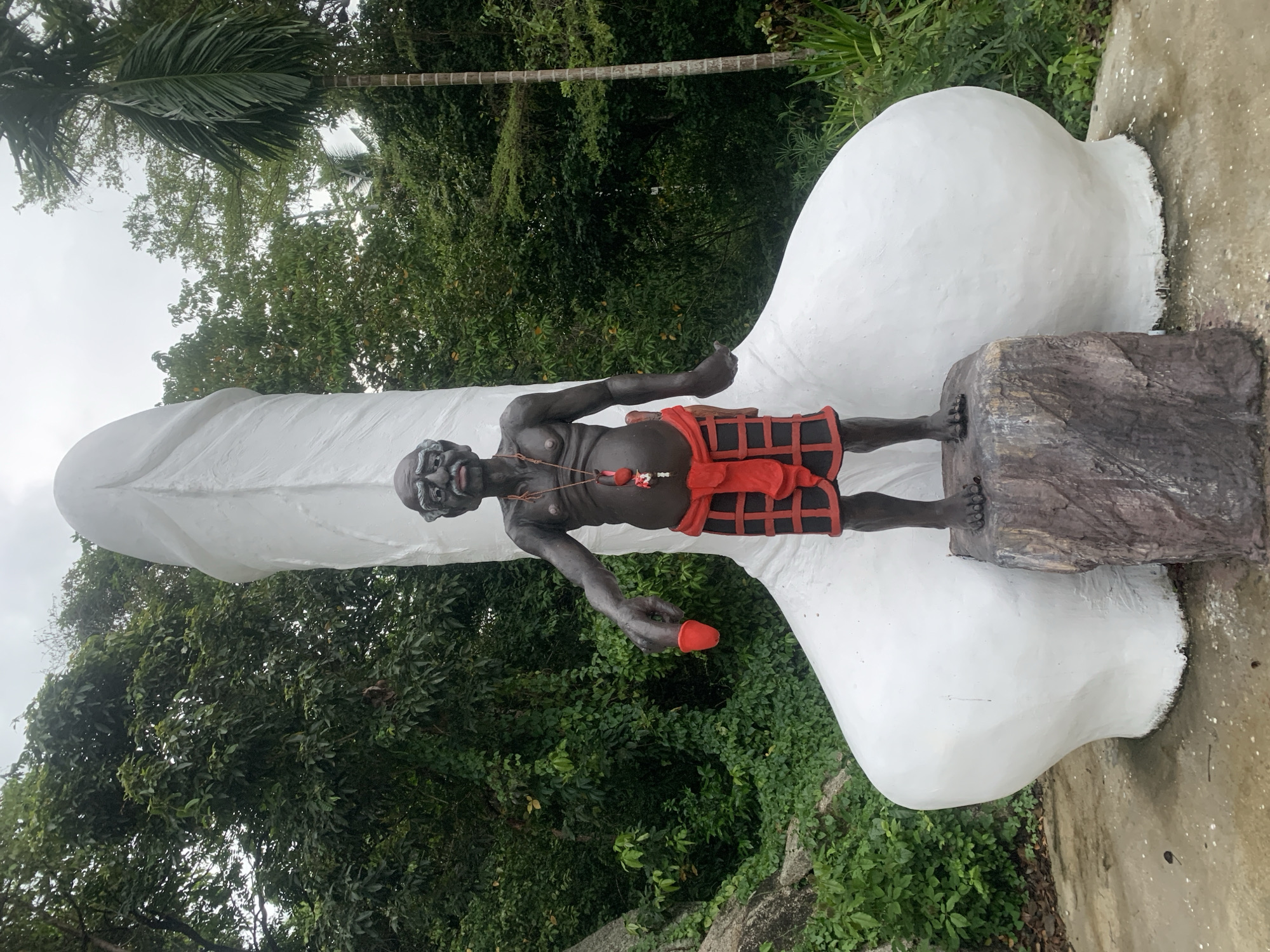 The image, presented rotated 90 degrees counter-clockwise from an upright orientation, depicts an outdoor scene featuring a dark-skinned statue of a man beside a prominent, large white phallic sculpture. Lush green tropical foliage and a bright, overcast sky form the background.
The dark-skinned statue of the man stands on a rough, rectangular pedestal, positioned on what appears to be the lower right side of the large white sculpture. He exhibits an aged appearance with distinct facial features and a serious expression. His bare chest reveals prominent ribs, and he wears a necklace of red beads with a larger red pendant. A red, plaid-patterned loincloth or skirt covers his waist. In his right hand, extended downwards, he holds a small, bright red, phallic-shaped object.
The substantial white sculpture dominates the frame, extending horizontally from the lower right to the upper left. Its smooth, rounded surface is uniformly white and distinctly phallic in form. The human statue is positioned near its base.
The background is filled with dense tropical trees and leafy vegetation, including a palm tree trunk and fronds, all appearing rotated due to the image's orientation. The sky, visible at the top and left, is bright and cloudy, indicating daytime. The immediate ground beneath the figures is a mix of concrete and natural earth. No text is visible in the image. This arrangement suggests a cultural or religious display, possibly a fertility shrine, characteristic of locations such as Baan Hua Thanon, Thailand.