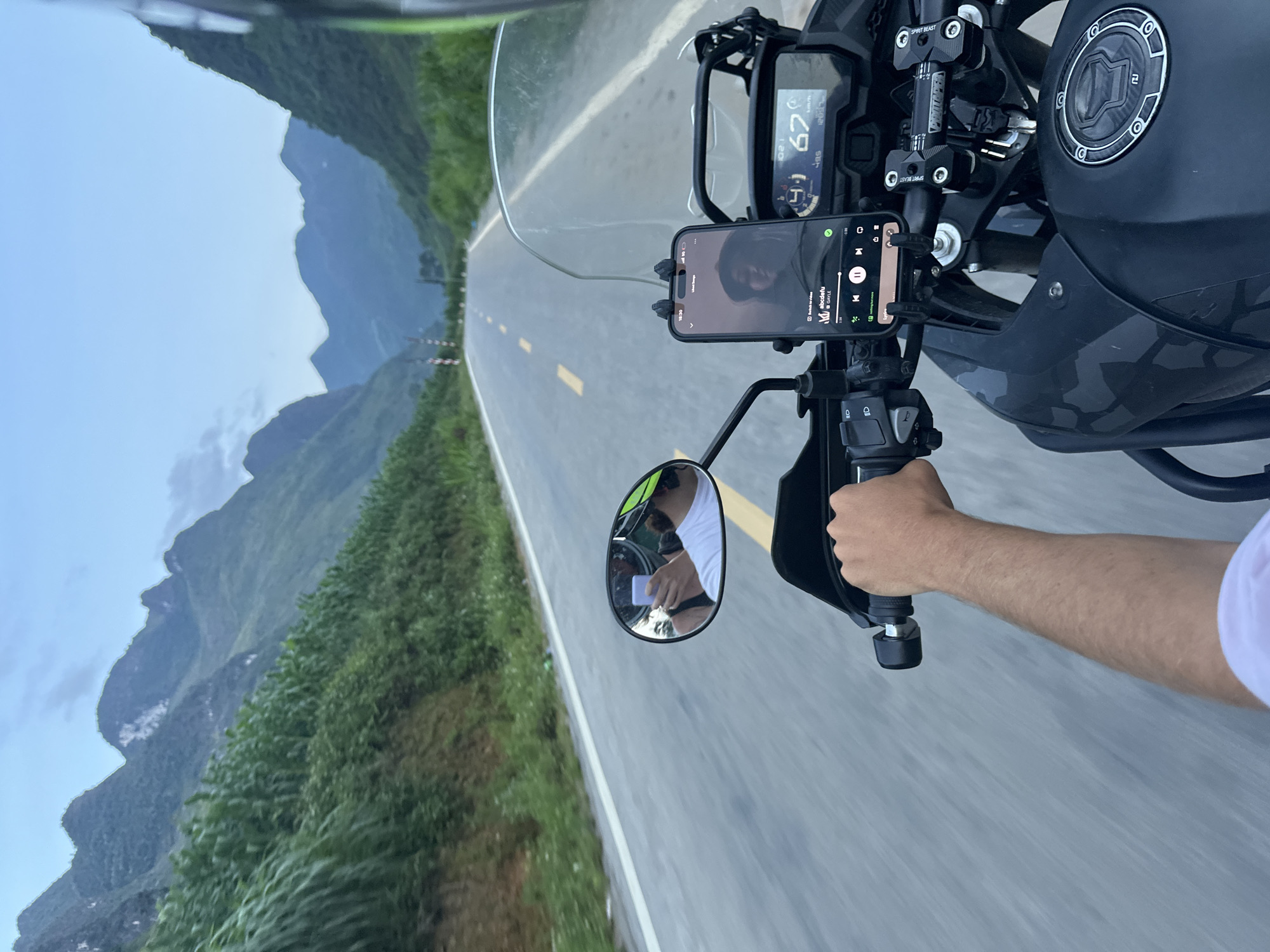 A person is riding a motorcycle on a paved road, visible from the perspective of the rider. The motorcycle has a digital speedometer reading 67 km/h and a smartphone mounted displaying a music app. A left-side mirror reflects the rider's arm and torso. The road is lined with vegetation, leading toward distant mountains under a blue sky. The location is Mèo Vạc Commune, Vietnam.