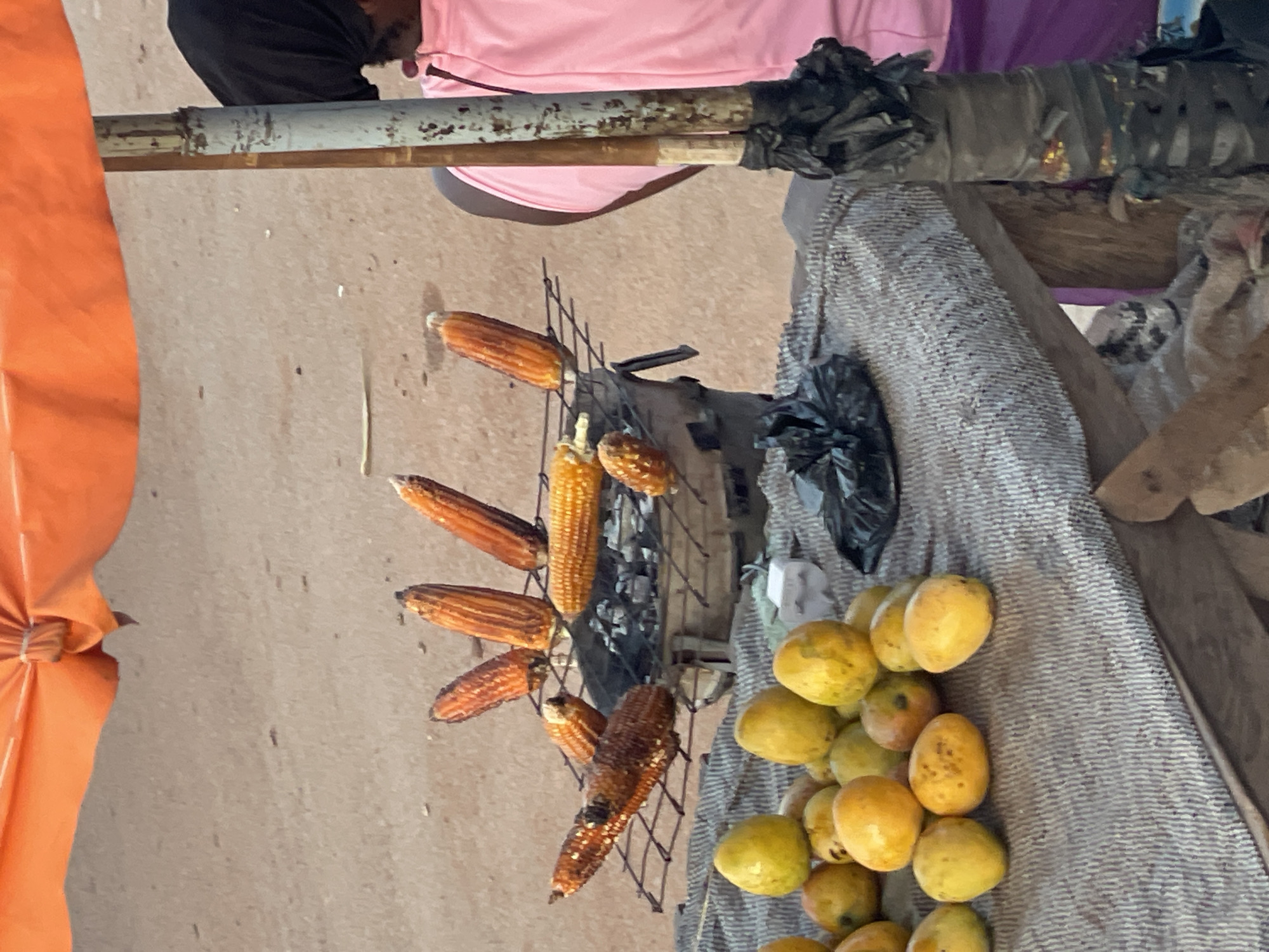 An overhead view shows a street vendor's stall. An orange awning is visible at the top of the frame, casting a shadow over the scene. Beneath the awning, a collection of grilled corn on the cob is arranged on a metal rack over a charcoal grill. The corn cobs are a deep orange-brown color, some appear to be split open. To the left of the grilled corn, a pile of yellow and green fruits, likely mangoes, are displayed on a gray, textured cloth. In the background, a person wearing a pink shirt and a dark head covering is visible behind the stall. The ground is a dusty, sandy surface. The scene appears to be set up outdoors, possibly in a market or on a street.