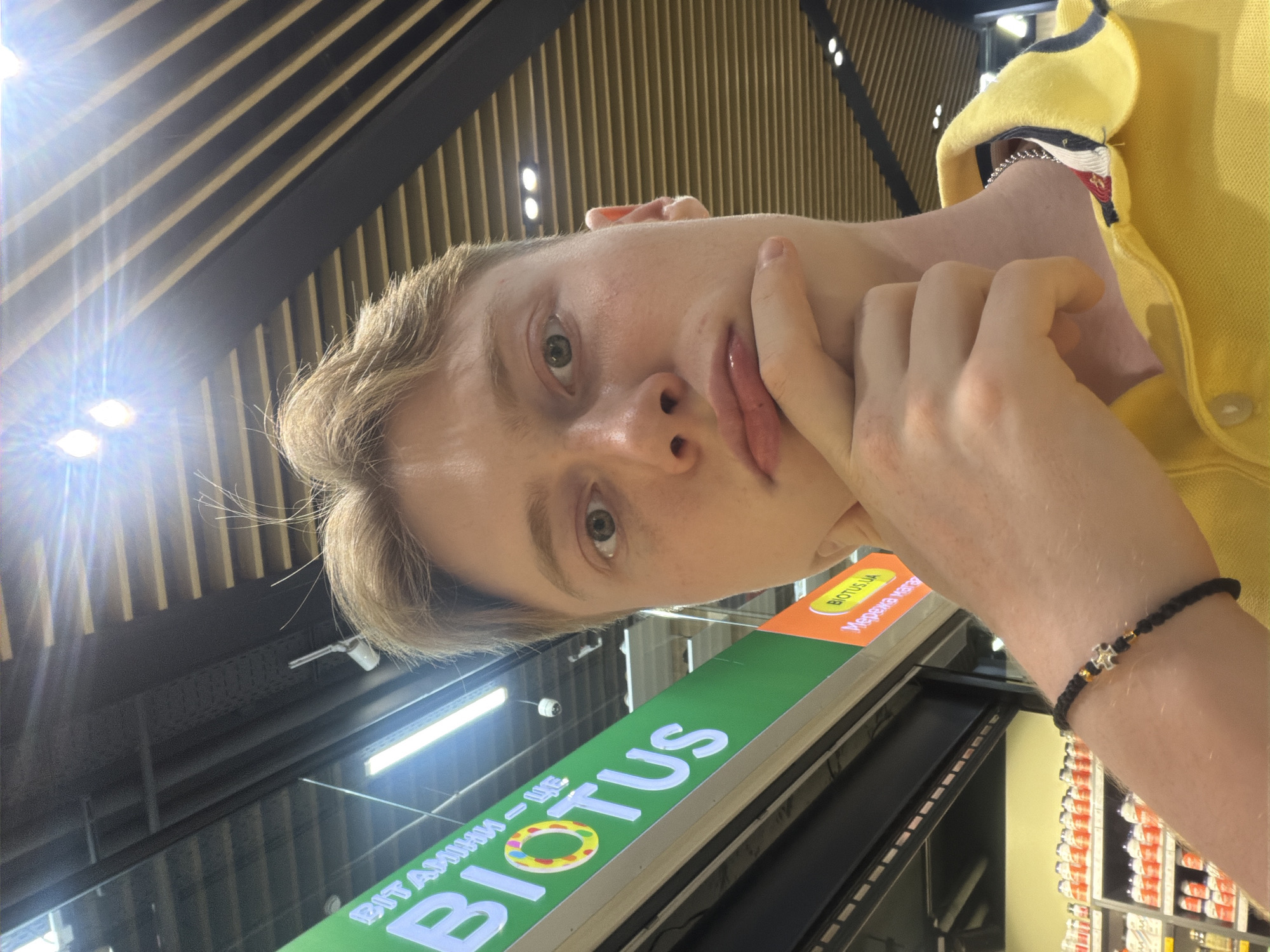 A young Caucasian male with light brown hair and green eyes is captured in a close-up, head-and-shoulders selfie, with his left hand resting on his chin. He is wearing a yellow polo shirt. The background indicates he is indoors, likely in a store, with bright fluorescent lighting and a slatted ceiling visible. A green sign with white text, reading "ВІТАМІНИ - ЦЕ BIOTUS" (Vitamins - This is BIOTUS), is prominent on the left side of the image, suggesting the location is a BIOTUS store. A smaller orange sign with "BIOTUS.UA" is also visible. The overall impression is a casual, indoor portrait taken with a smartphone.