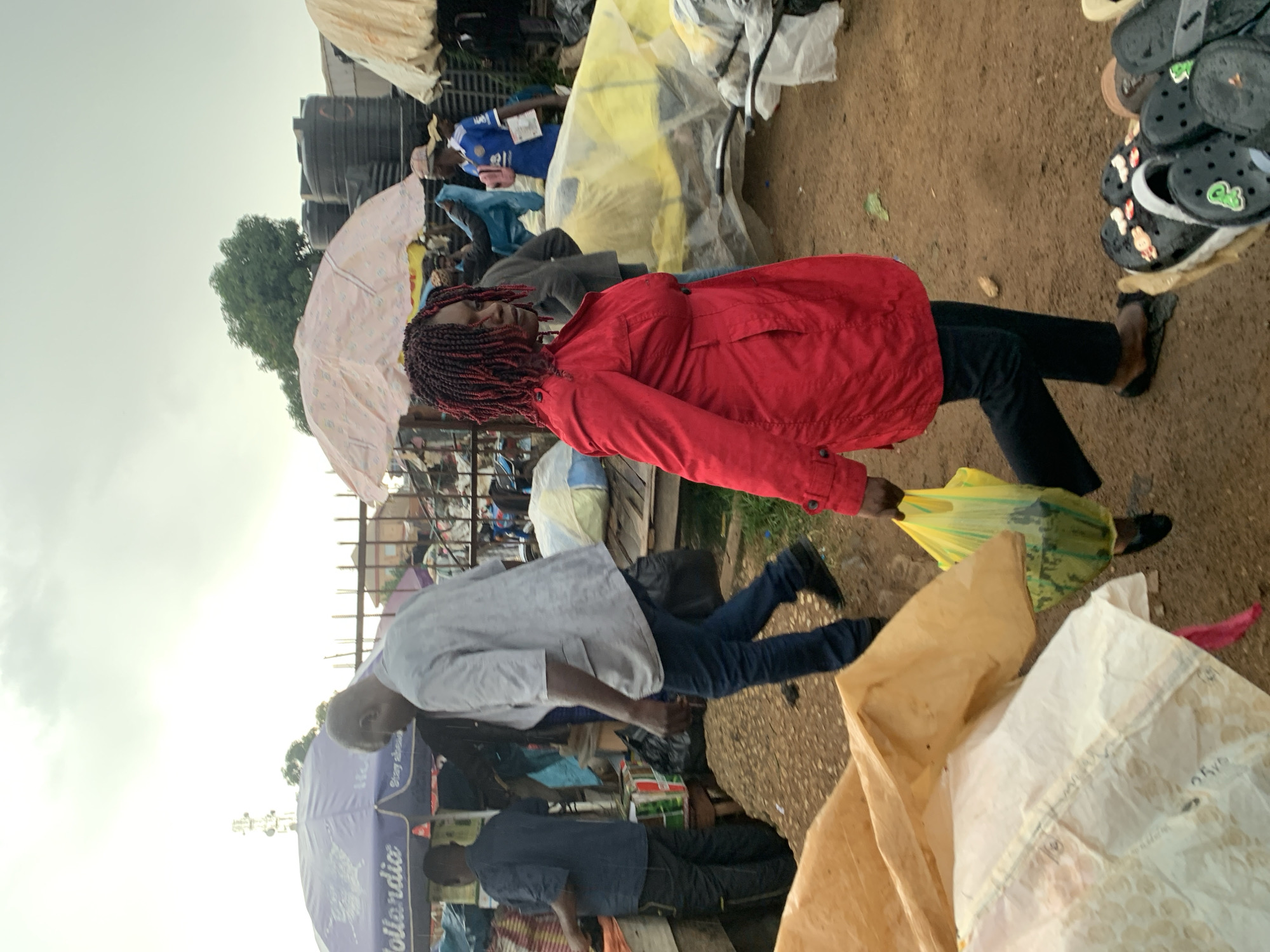 In Gura Top, Nigeria, a scene unfolds at an outdoor market under an overcast sky. A woman with braided red and black hair, wearing a red jacket and black pants, walks from right to left, carrying a yellow plastic bag. She is in the foreground, facing the left side of the frame, her gaze directed downwards. Behind her, other individuals navigate the market. A man in a light gray jacket and blue jeans walks in the opposite direction, carrying a black bag. Another man, wearing a blue shirt, walks towards the background. Umbrellas, some colorful and some plain, provide shade over stalls. Large black water tanks are visible in the background, along with assorted goods and structures that suggest a bustling marketplace. The ground is dusty and uneven, with piles of goods and debris scattered around. In the lower right corner, a display of various footwear, primarily black clogs and sandals, is visible.