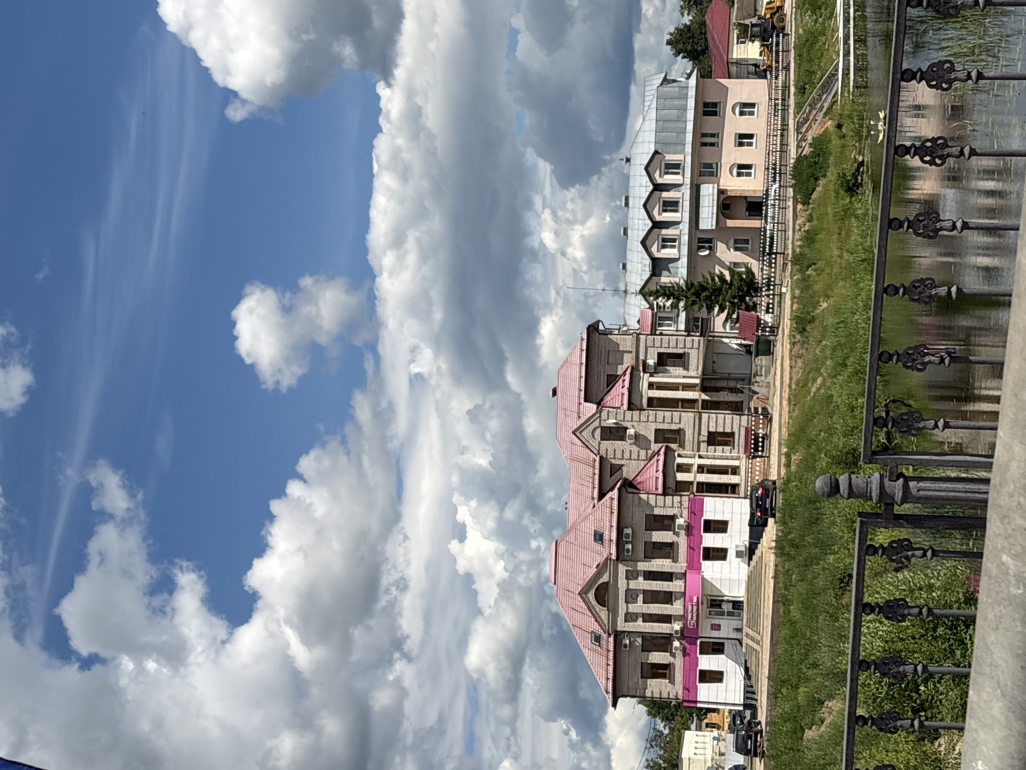 This image depicts a view of buildings in Pereslavl-Zalessky, Russia, situated along a waterfront. In the foreground, a dark metal fence with intricate scrollwork runs horizontally across the lower portion of the frame, partially obscuring a reflective body of water. The water's surface shows ripples and reflections of the surrounding environment.
Behind the fence, a grassy embankment slopes upwards towards a row of buildings. The buildings are of varying architectural styles. The building on the left is a multi-story structure with a light-colored brick facade and a prominent pink tiled roof. It features numerous windows and some visible air conditioning units. A pink stripe with text, likely signage, runs along the lower portion of this building. Adjacent to it is another building, also multi-story, with a beige facade and a silver-colored pitched roof with multiple dormer windows. This building has a more modern appearance. Further to the right, another building with a red roof is visible, along with part of a yellow construction vehicle.
The sky above is a bright blue, dotted with large, fluffy white cumulus clouds, creating a dynamic and somewhat dramatic contrast with the scene below. A few distant trees are visible beyond the buildings, suggesting a semi-urban or suburban setting. The overall impression is that of a sunny day with dramatic cloud formations over a town by the water.