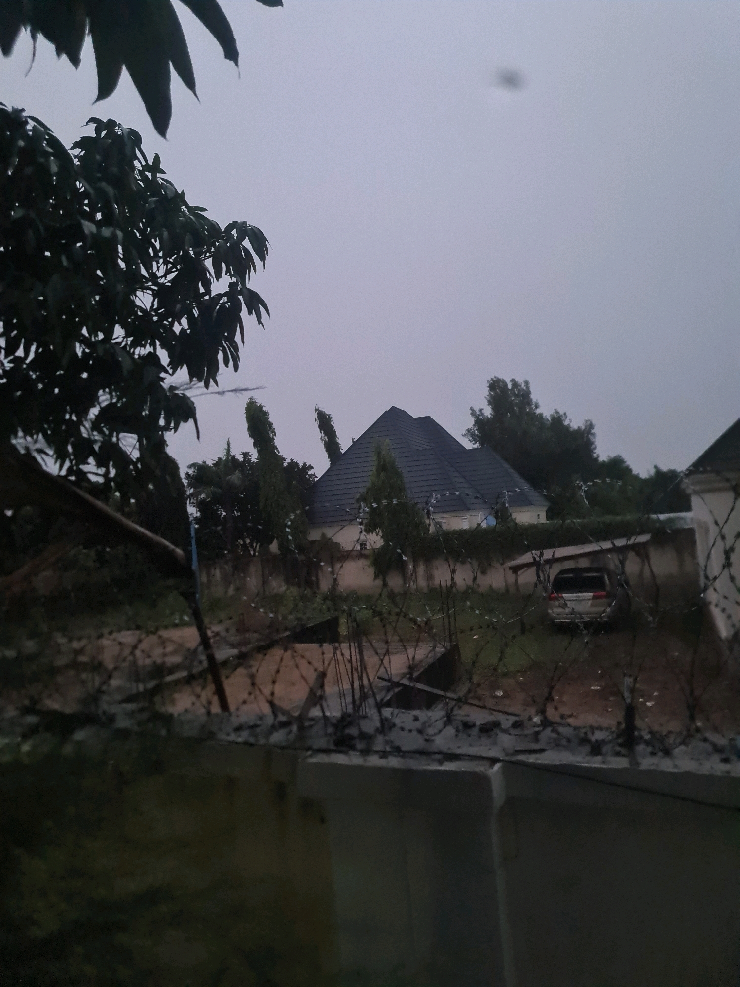 The image captures a cloudy, overcast day in Wauro Jabbe, Nigeria. In the foreground, a concrete wall and barbed wire fence are visible, suggesting a boundary or security measure. Beyond the fence, a residential area is depicted. A modern house with a dark, angular roof is prominent, flanked by lush green trees. To the right, another building, partially visible, also appears to be part of the same property. A silver minivan is parked under a rudimentary shelter, its rear facing the viewer. The overall scene is one of a residential compound under a somber sky.
