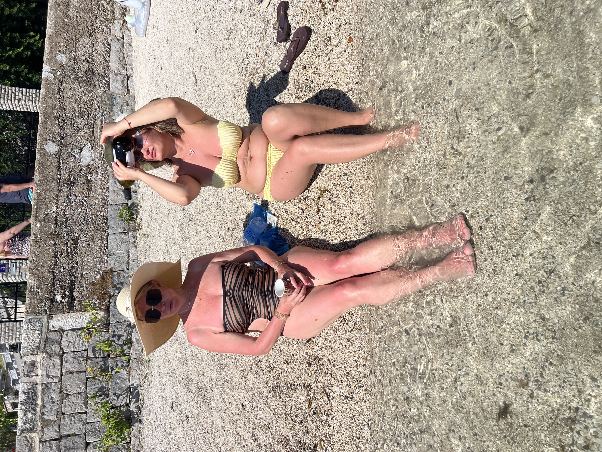 Two women are sitting on a pebble beach in Đenovići, Montenegro, with their legs in the shallow, clear water. The woman on the left, wearing a striped one-piece swimsuit and a wide-brimmed hat, is holding a cup and has sunburnt shoulders and legs. The woman on the right, wearing a yellow bikini, has a bottle of wine on her head and is holding her hat. Both women are wearing sunglasses. The background shows a stone wall and some vegetation. There are also flip-flops on the beach near the woman on the right.
