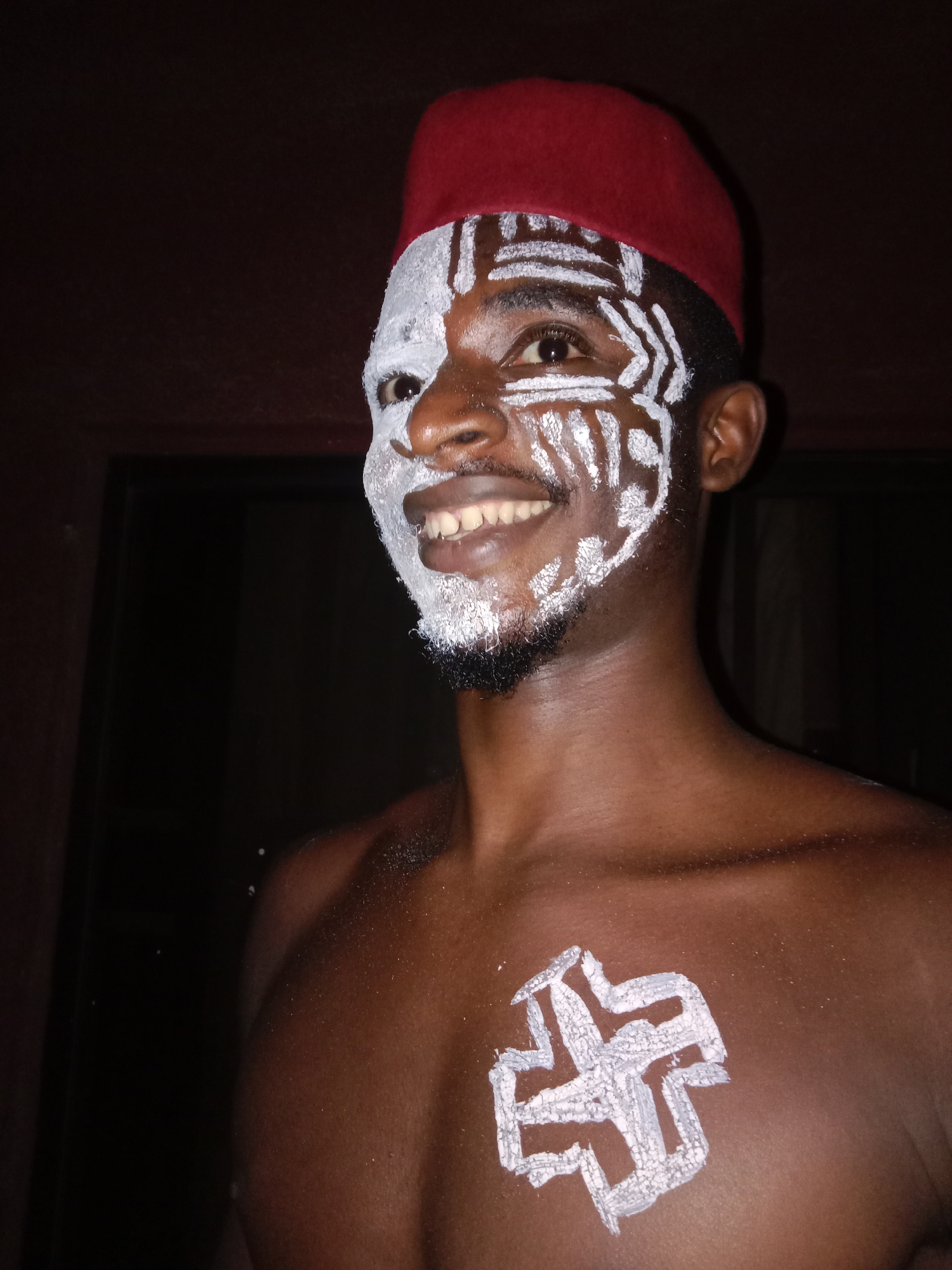 A man is depicted from the chest up. He is wearing a red fez and has white paint on the left half of his face in patterned lines. A white symbol is drawn on his chest. The background is dark. The location is Unknown, Unknown.