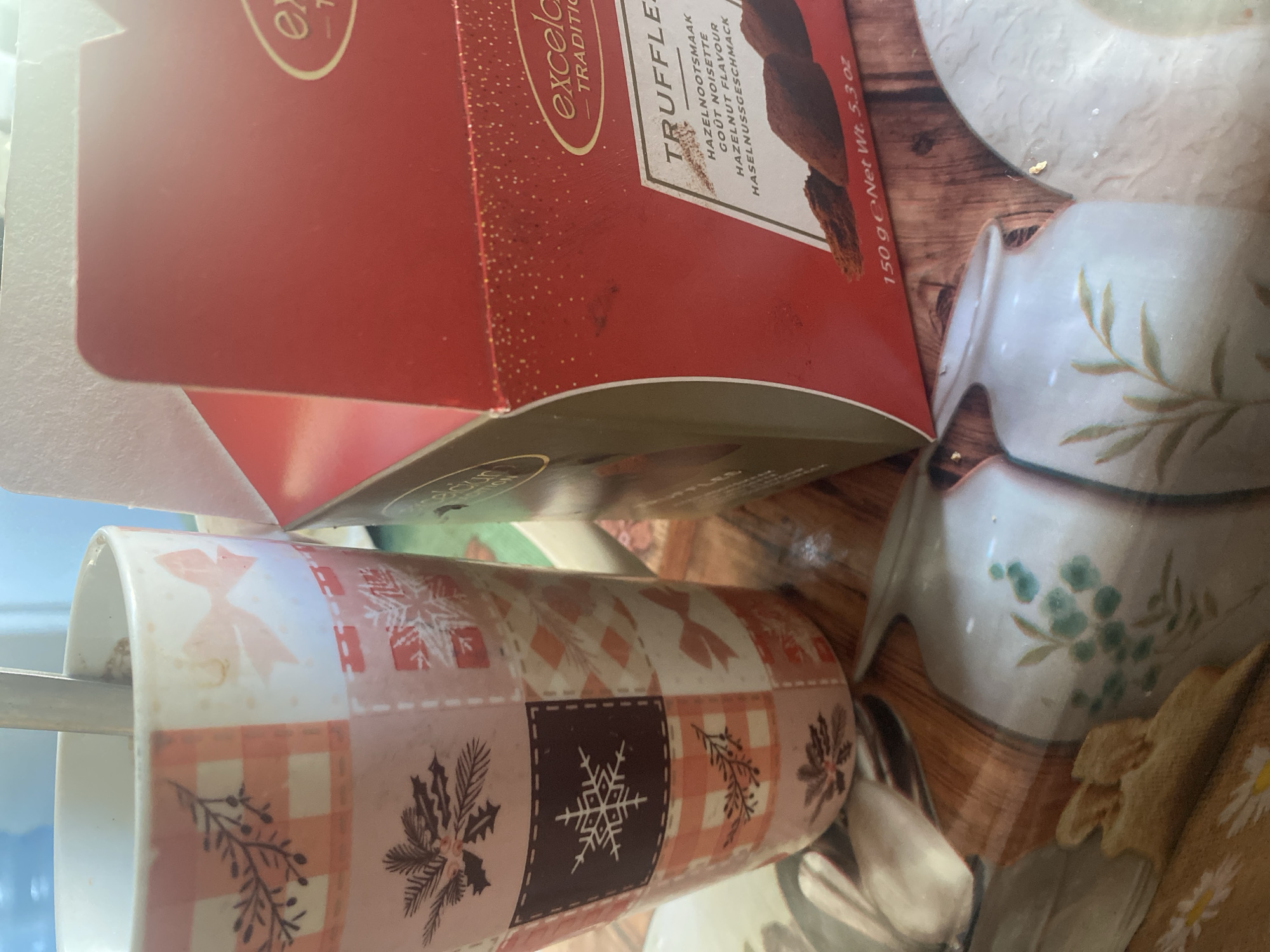 The image captures a close-up view of a table setting, likely indoors. In the foreground, a white mug with a patchwork design featuring orange checks, snowflakes, holly sprigs, and bows is positioned on the left. A metal spoon is visible inside the mug. To the right of the mug is an open red box of "excelcior TRADITION" truffles. The box is adorned with gold speckles and features text indicating "TRUFFLES," "HAZELNOOTSMAAK," "GOUT NOISETTE," "HAZELNUT FLAVOUR," and "HASSELNUSSGESCHMACK." Below this text, a few dark brown truffles are visible. The background is blurred, but appears to show a wooden surface with some decorative elements, possibly ceramic figurines with floral patterns. The lighting suggests an indoor environment, likely during the daytime. There is no visible indication of the location being in Dnipro, Ukraine, or any specific city.