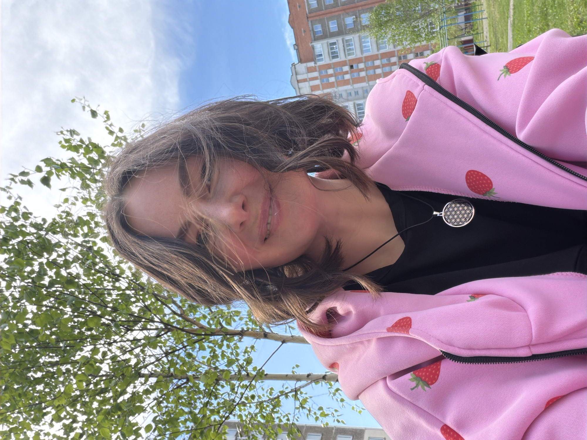 A young person poses for an eye-level selfie outdoors. The subject, facing towards the camera at an angle, is wearing a light pink hooded sweatshirt with a strawberry pattern over a black shirt, accented with a silver pendant necklace. The sky, tinted with light blue and scattered clouds, forms the backdrop. Elements of an urban scene are also present, including trees with fresh green leaves, and apartment buildings in the distance. The setting appears to be a park or recreational area within a city.