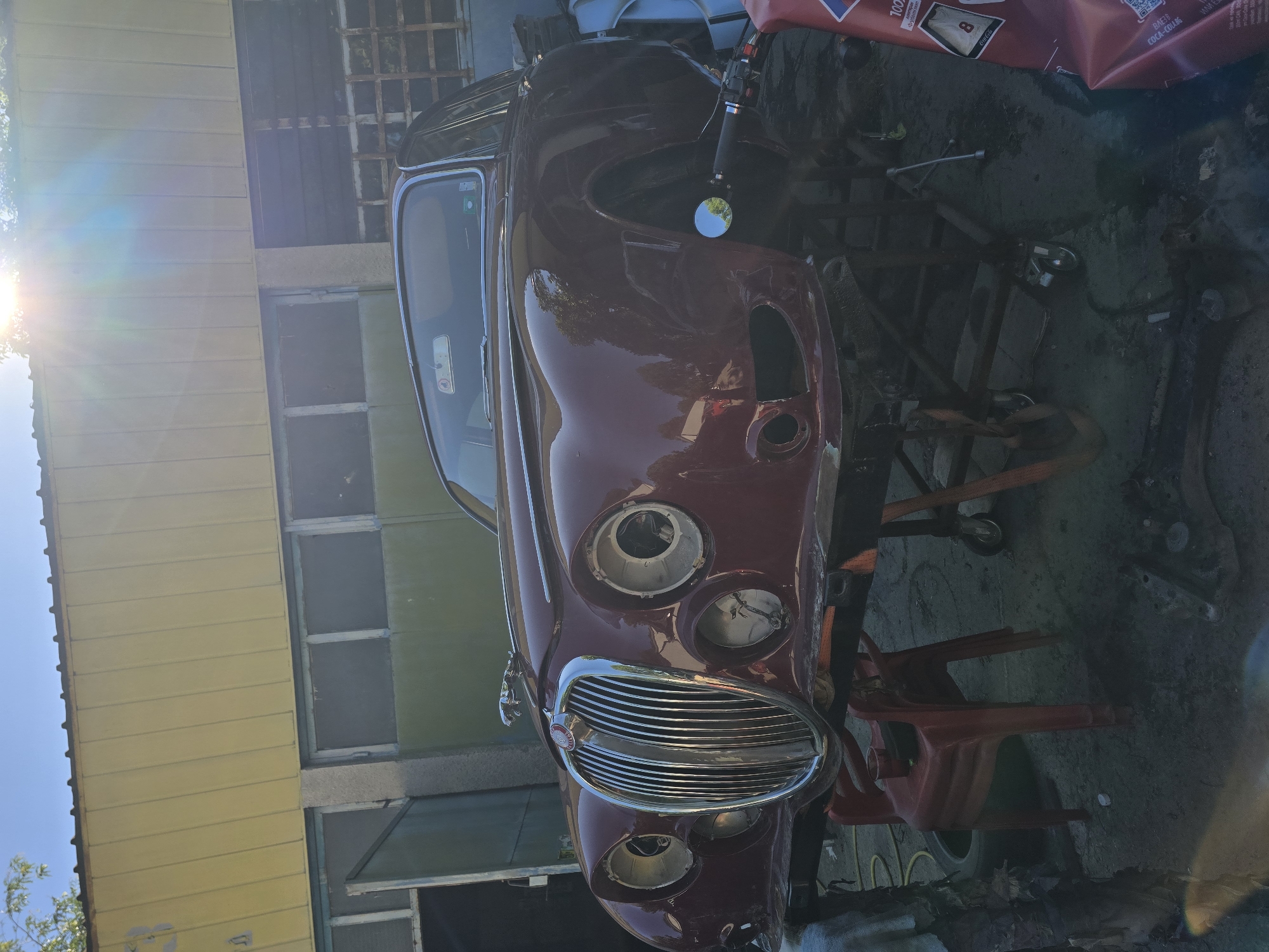 An aged, maroon Jaguar Mk2 sedan is undergoing restoration in an outdoor workshop or yard. The car is elevated on a stand, with its front grille and headlight assemblies prominently displayed. The car appears to be missing some of its exterior components, such as headlights and possibly the front bumper. A portion of a yellow building with a corrugated metal roof is visible in the background, along with some windows and what appears to be a garage door. The sun is shining brightly, casting lens flare on the image. The ground is concrete, with some scattered tools, tires, and other debris. The setting is likely an industrial or automotive repair area, possibly in Ruse, Bulgaria, given the context. There are no people visible in the image.