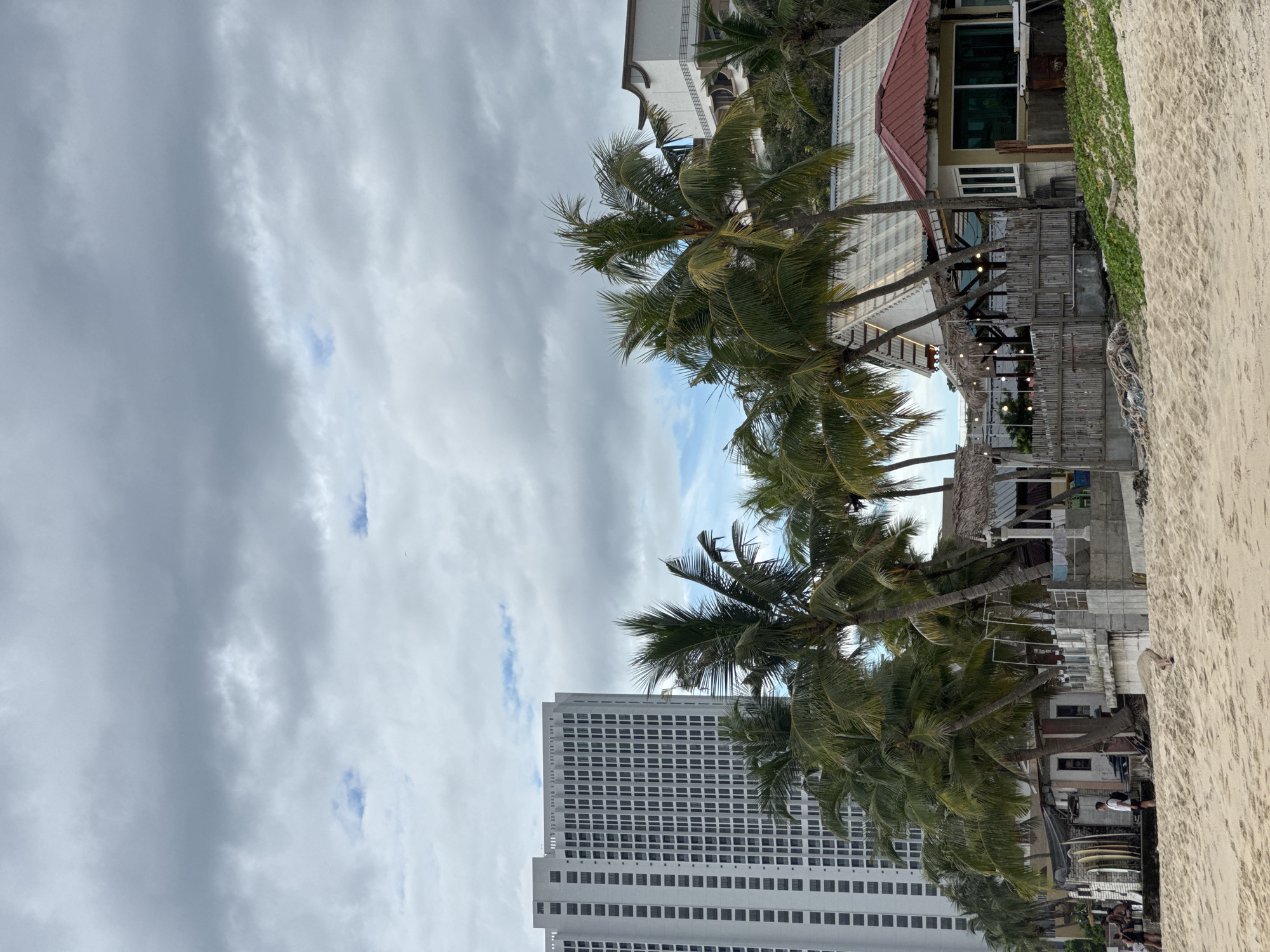 A wide shot captures a beach scene with a towering white apartment building on the left, several palm trees in the mid-ground, and small beachside structures in the foreground. The sky is overcast with heavy gray clouds. The beach is composed of light-colored sand.
To the left of the frame, the apartment building features a repetitive pattern of dark windows. In front of it and extending towards the center, are several mature palm trees with lush green fronds, some leaning at angles. Behind the palm trees, several small, rustic buildings with thatched roofs and wooden structures are visible, some appear to be shops or cafes. One structure on the far right has a red roof and green-tinted windows.
In the far left foreground, a few people are visible on the sand, appearing to be walking or standing. One person is standing near a collection of surfboards leaning against a wall. Another structure, made of concrete blocks and posts, is situated behind the sand and in front of the beachside huts. Some laundry is hanging on a clothesline on this structure.
The overall scene suggests a tropical beach destination with a mix of modern development (the apartment building) and traditional beachside amenities.