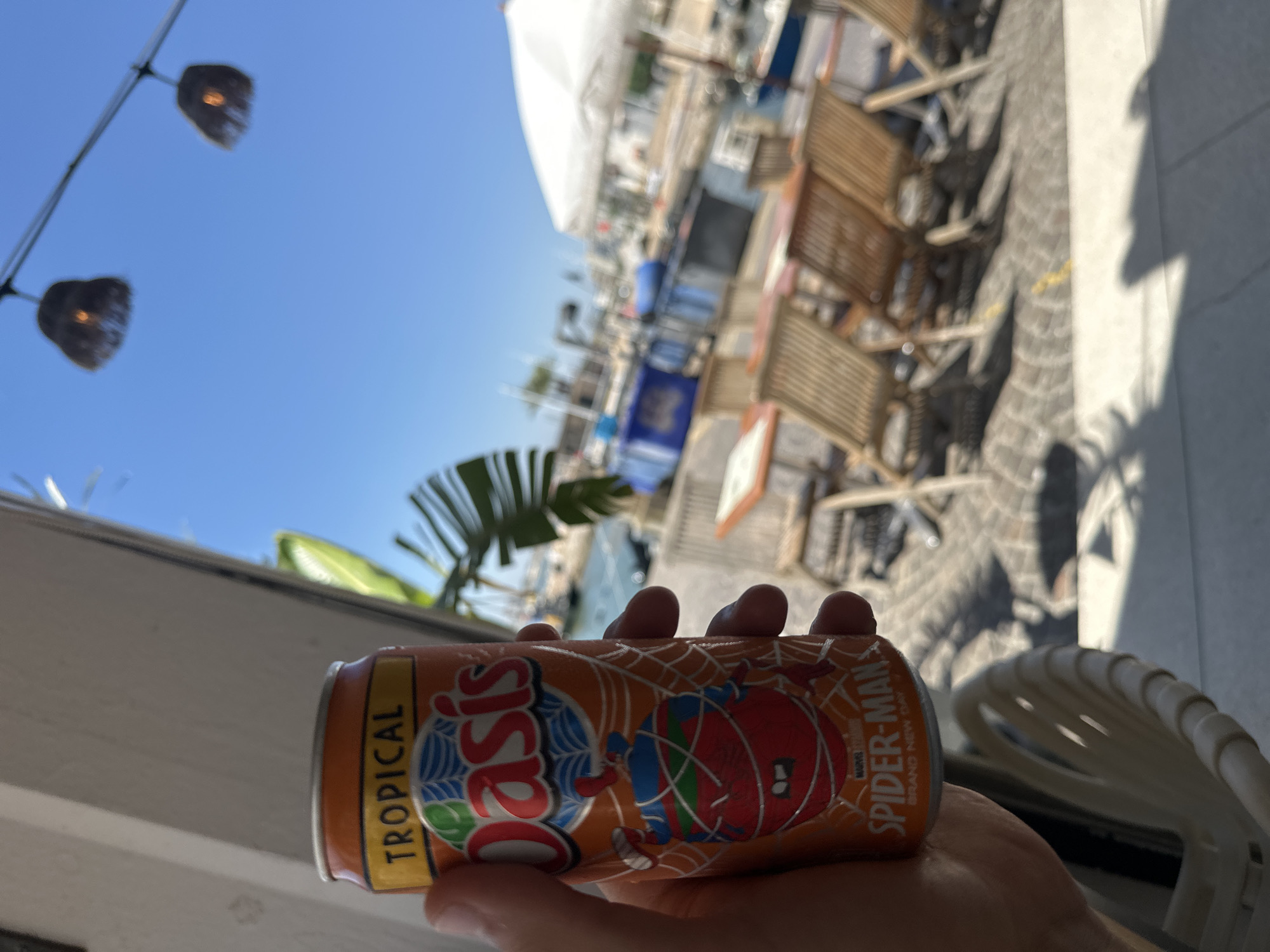 A person's hand holds an orange can of Oasis Tropical Spider-Man branded juice. The can features a spider web design and a cartoon image of Spider-Man. The setting appears to be outdoors, possibly a cafe or patio, with wooden chairs and tables visible in the blurred background. The sky is bright blue, suggesting it is daytime, likely sunny. There are also some tropical plants in the background. The lighting indicates a sunny day. The focus is on the can of juice. Visible text on the can includes "Oasis," "TROPICAL," and "SPIDER-MAN."