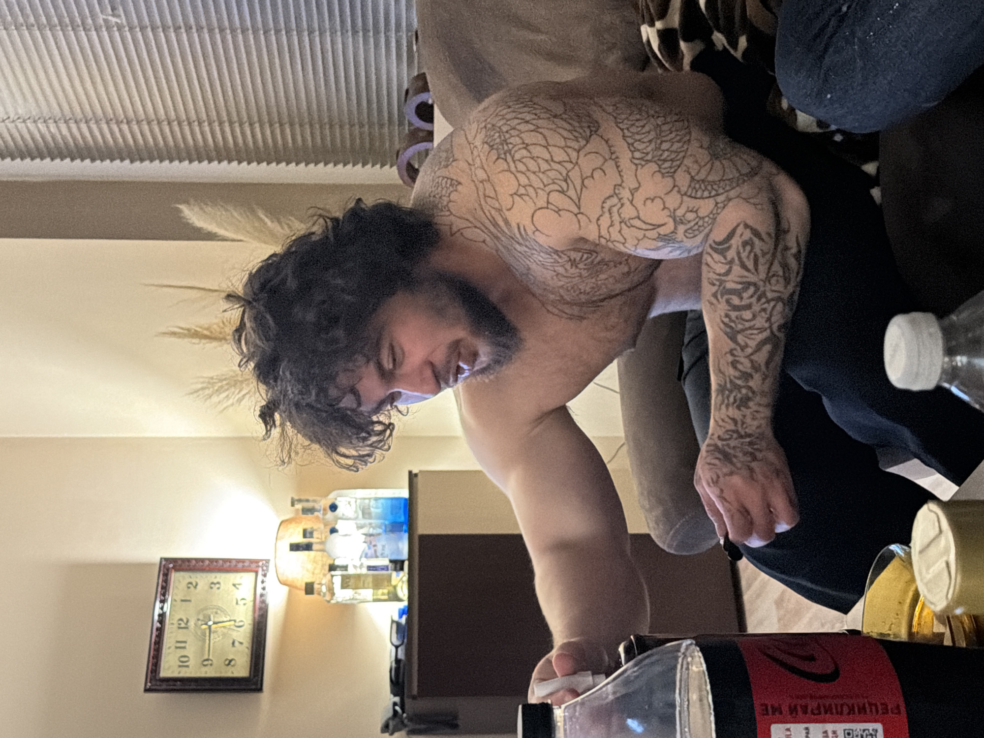 A man with dark, curly hair and tattoos on his arms and back is leaning over, holding a small white object, possibly a pill or a piece of paper. He appears to be looking down at something on a surface in front of him. The environment includes a room interior with a clock on the wall, various bottles on a shelf, and window blinds in the background. The exact city of Unknown, Unknown is not discernible from the image.