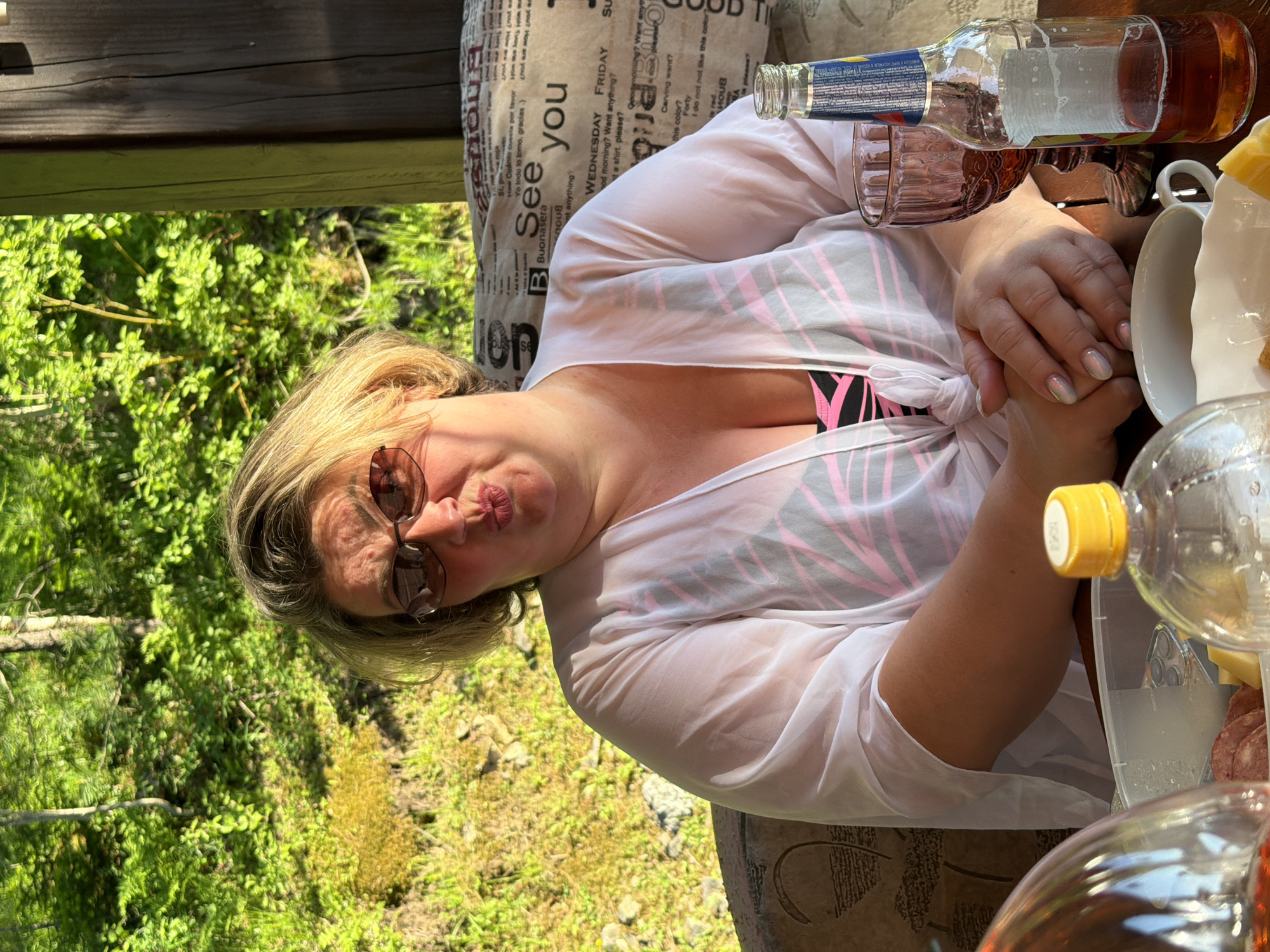 A middle-aged Caucasian woman with blonde hair and sunglasses is seated at a table outdoors. She is wearing a white sheer cover-up over a pink and black swimsuit. Her hands are clasped in her lap. A bottle of beer, a glass, and a plastic container with food are on the table in front of her. The background shows a lush green forest. The setting is likely a dacha or a picnic area near Tashtagol, Russia, given the vegetation and style of wooden structure visible.
