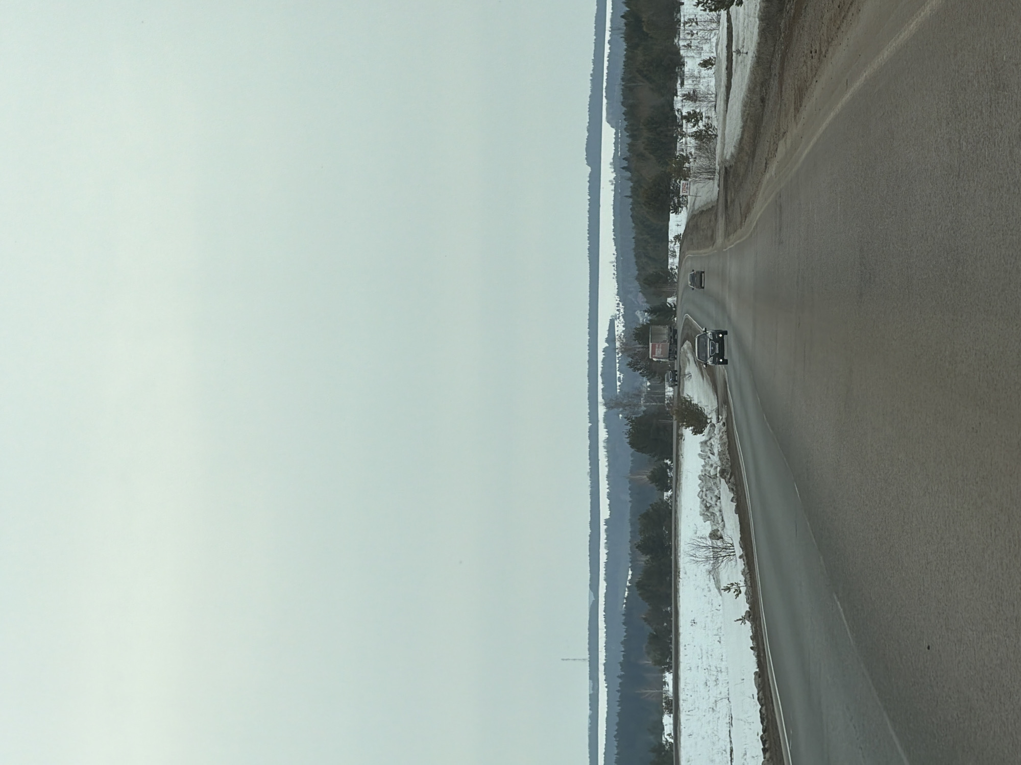 The scene depicts a two-lane asphalt highway extending into the distance within the vicinity of Povarenki, Russia. The sky is uniformly overcast. The road surface is dark, appearing damp or worn, with a single dashed white line marking the center. Snow-covered ground with patches of exposed earth and sparse vegetation, including small coniferous and bare deciduous trees, borders both sides of the highway. A dense line of dark coniferous trees forms a midground horizon. Beyond this, a wide, flat expanse of white, consistent with snow-covered land or a frozen body of water, stretches across the background. A faint, tall, slender, dark tower structure is visible on the left side, rising from this white expanse. A distant, dark tree line marks the far horizon. Four vehicles are observed traveling away from the viewer: a dark sedan in the foreground, a lighter-colored sedan further ahead in the same lane, a white semi-truck with a red cab and red lettering on its trailer positioned in the left mid-distance, and another lighter-colored car on the right side of the road in the mid-distance.