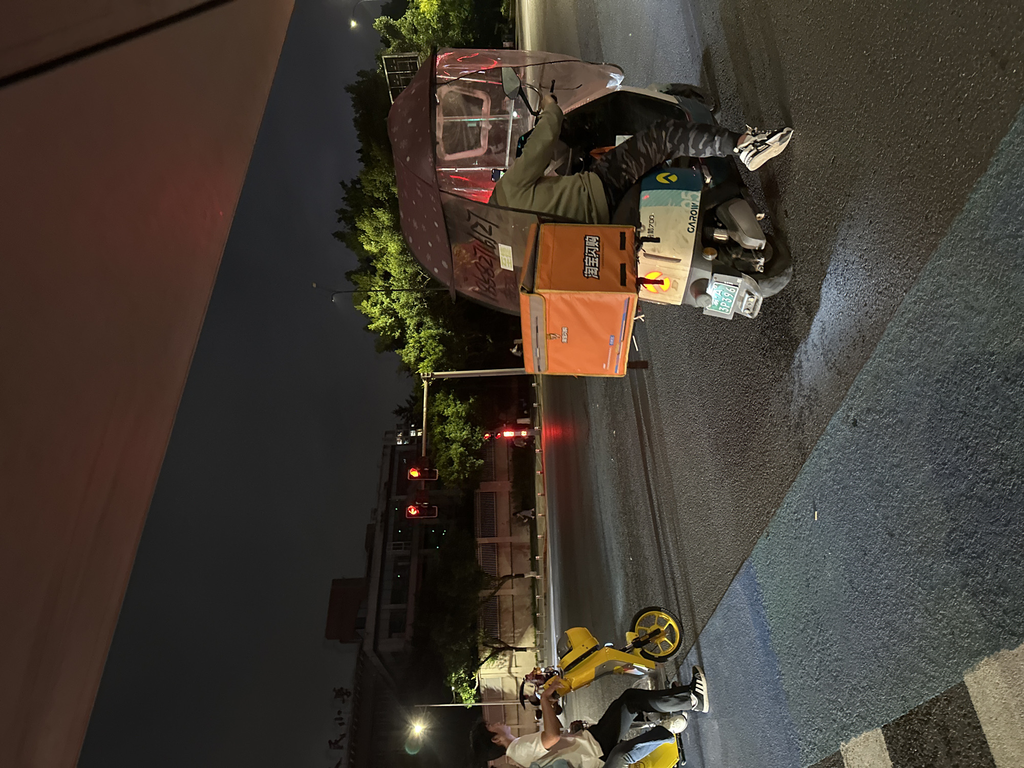 At night, a person is riding a scooter with a large orange insulated box attached to the back in the Chongqing High-tech Zone, China. The scooter has a clear plastic enclosure with a patterned roof, providing some protection from the elements. The rider is wearing camouflage pants and a grey long-sleeved shirt. Another scooter, yellow in color, is visible to the left, with two people on it. The street is paved and appears to be a road with traffic lights in the background, which are illuminated red. Buildings and trees are visible in the background, illuminated by streetlights. A pedestrian crossing with blue and grey markings is in the foreground.