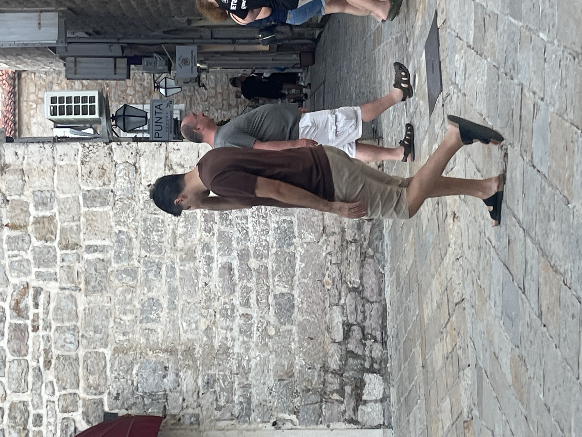 In Budva, Montenegro, a narrow, cobbled street runs between old stone buildings. A man in a brown t-shirt and beige shorts walks away from the camera, with his hand near his face. To his right, another man in a grey t-shirt and white shorts walks in the same direction. In the background, a few other figures are visible, along with architectural elements like a white air conditioning unit and a black lantern with a "PUNTA" sign. The stone walls and paving suggest an old European town.