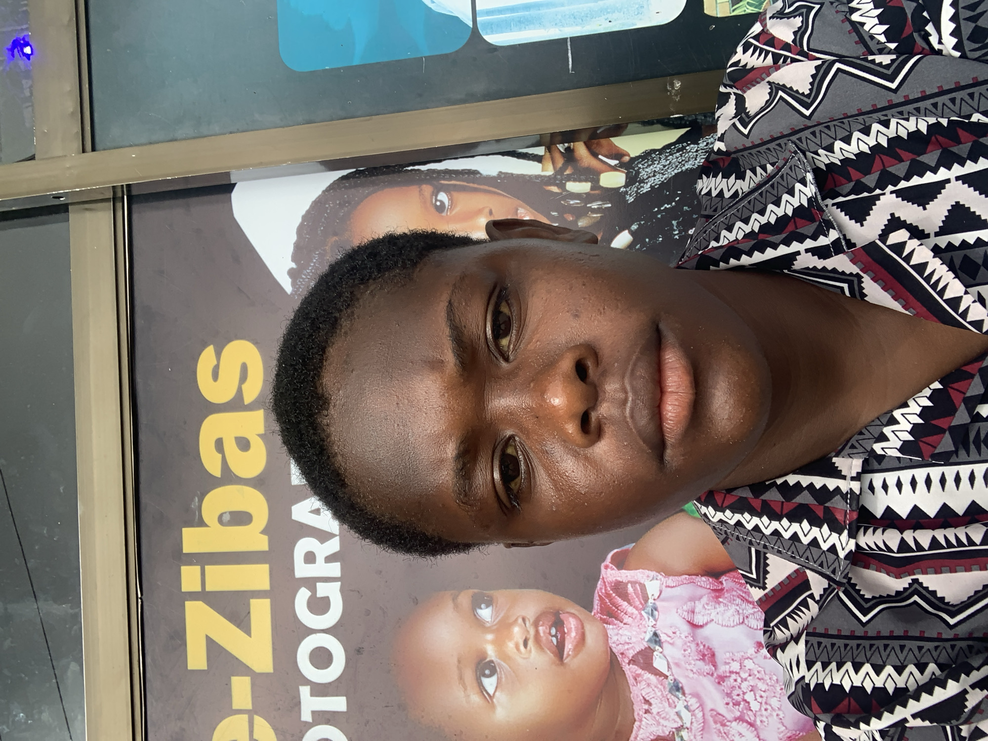 The image is a close-up, waist-up, selfie-style photograph of a young Black individual wearing a black, white, and maroon geometric-patterned shirt. The individual's head is turned slightly to the right, and they are looking directly at the camera with a neutral expression. Their skin appears slightly oily, and there are visible pores and some blemishes. The background features a large poster for "e-Zibas PHOTOGRAPHY," with images of children visible behind the individual. The poster is mounted on a building with a metallic frame, suggesting an outdoor setting, possibly a storefront or a public area in Jos, Nigeria. The lighting is even, indicating it was taken during the day.