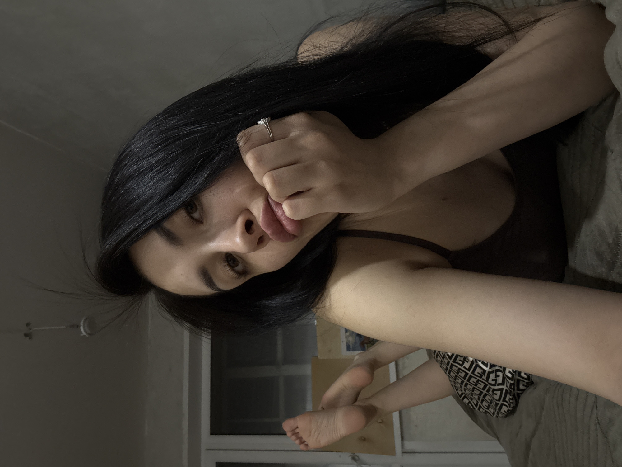 A woman with long, dark hair is lying down, partially obscured by a gray blanket. She is wearing a dark tank top and patterned shorts. Her face is close to the camera, and she is sticking her tongue out slightly. A ring is visible on her finger. A window with white frames and a wall can be seen in the background. The image does not provide information about the location in the city of Votkinsk, Russia.