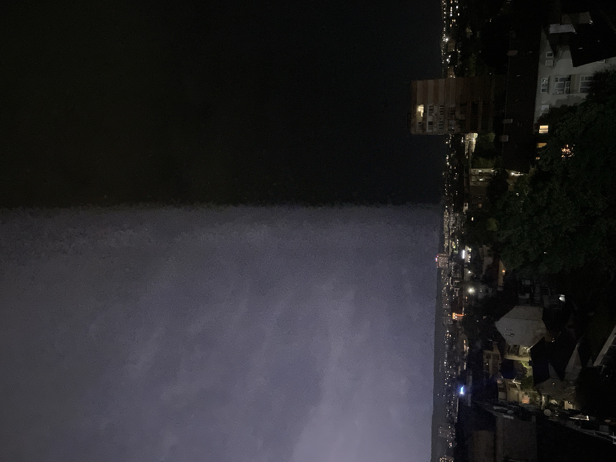 The image shows a cityscape at night, likely in an urban environment of Unknown, Unknown. On the right side, there are closely-packed buildings of varying heights, interspersed with trees and illuminated by numerous lights. The left side of the image is considerably darker, indicating a body of water, possibly a river or bay, or a dark sky. The environment is mostly still. The lack of people suggests a quiet, nocturnal scene.