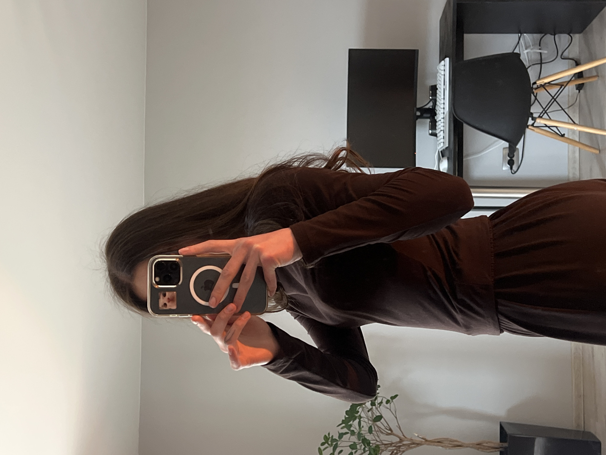 A person is captured in a mirror selfie, seen from the mid-chest down, obscuring their face with a smartphone. They have long, dark brown hair and are wearing a dark, possibly brown or deep maroon, long-sleeved top and matching skirt or wide-leg trousers, which appear to be made of a soft fabric. The top is tucked into the bottom garment.
The person holds a smartphone with a clear case that features a prominent silver MagSafe ring. A small picture or sticker of a light brown cat is visible on the phone case near the triple camera module.
The setting is an indoor room, likely a home office or bedroom. In the background, on the right side, there is a black desk with a black computer monitor and a white keyboard. A black office chair with wooden legs is pushed under the desk, and various cables are visible behind it. On the left side, a plant with several twisted stems stands in a black pot on the floor. The walls are a light, neutral color.
The image captures a casual moment of self-documentation. No specific time of day is discernible, with the indoor lighting providing illumination. No distinct text is visible, although the MagSafe ring implies an Apple device. The location context for this indoor scene is Ternopil, Ukraine.