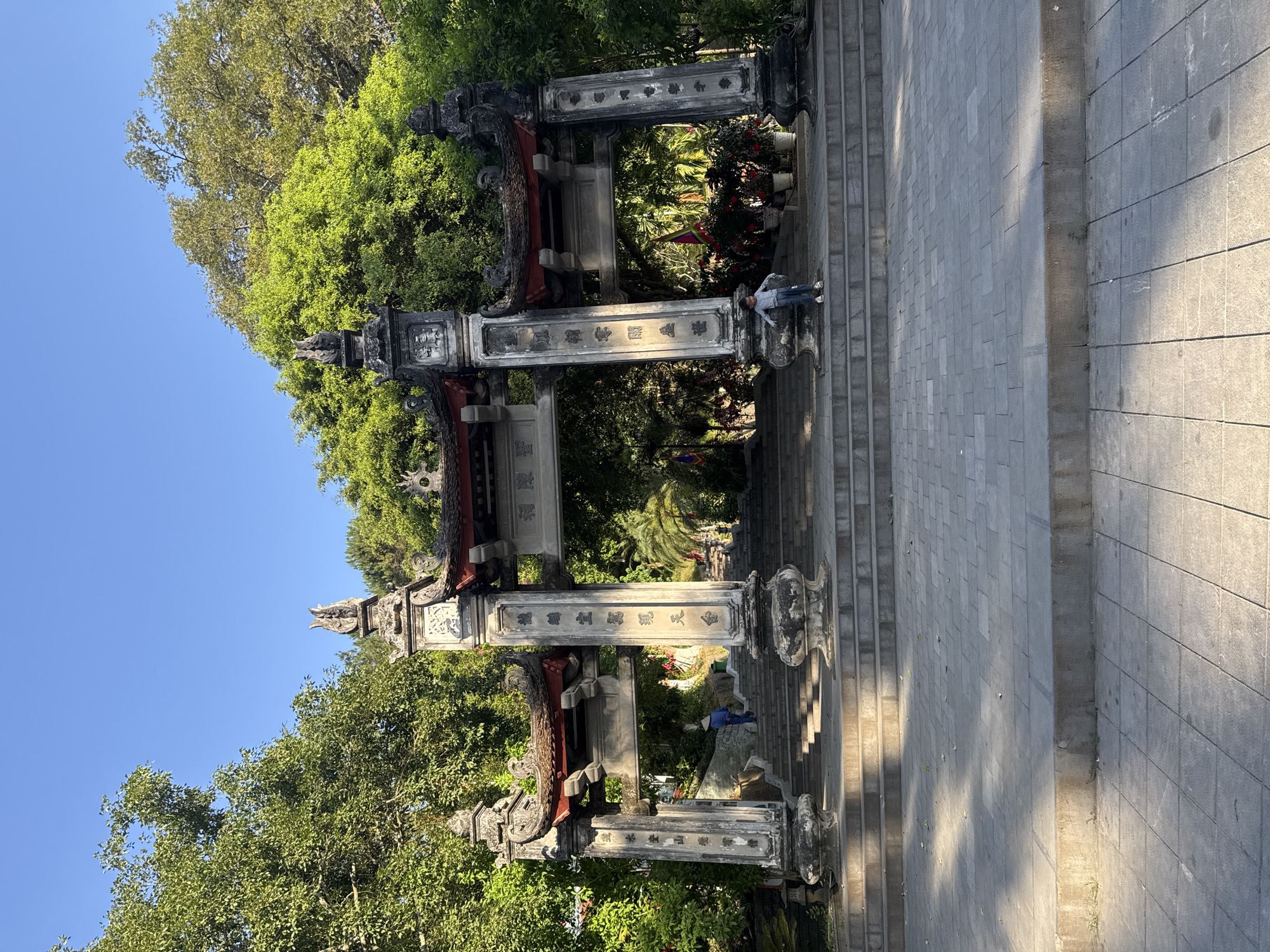 The image shows an ornate, stone entrance gate situated in Lao Cai Ward, Vietnam, with a steep staircase leading up to it. The gate consists of multiple pillars adorned with carvings and text, framed by lush greenery and blue sky. Two people stand within and behind the gate, one near the top of the staircase and the other further down. The staircase is made of rectangular stone blocks, with the paved ground extending to the right. The scene is bathed in sunlight.