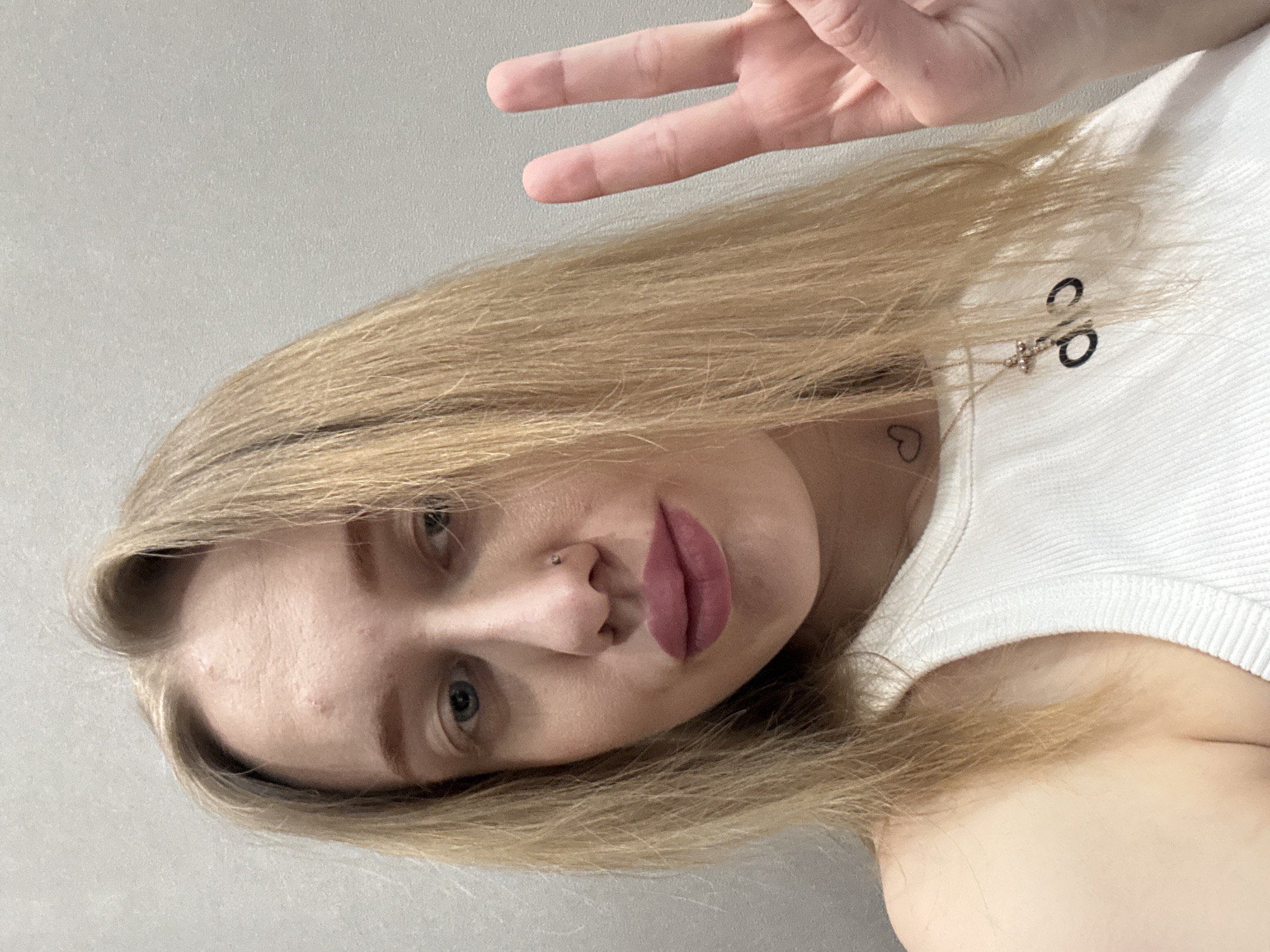 A fair-skinned woman with blonde hair is posing for a selfie. She is wearing a white tank top and has a nose ring. Her left hand is raised, forming a peace sign. A delicate necklace adorns her neck, and a small heart tattoo is visible on her upper chest. The background is a plain, light gray wall. The location is not discernable from the image.