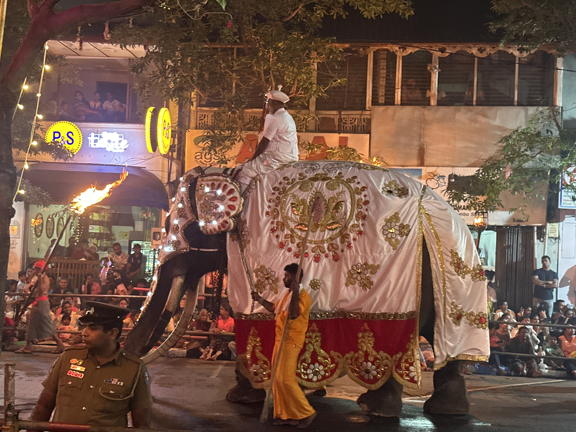 This image captures a nighttime procession in Kandy, Sri Lanka, likely part of a religious festival or parade. A large, elaborately decorated elephant is the central focus, draped in a white cloth adorned with gold embroidery and red trim. A man in a white shirt and hat sits atop the elephant. Another man, dressed in a bright yellow garment, walks alongside the elephant, holding a stick. In the foreground, a police officer in uniform stands watch. The background reveals a street scene with buildings, trees, and a crowd of onlookers gathered to witness the event. Streetlights and decorative lighting illuminate the scene, casting a warm glow. The architecture of the buildings suggests a traditional South Asian urban setting.