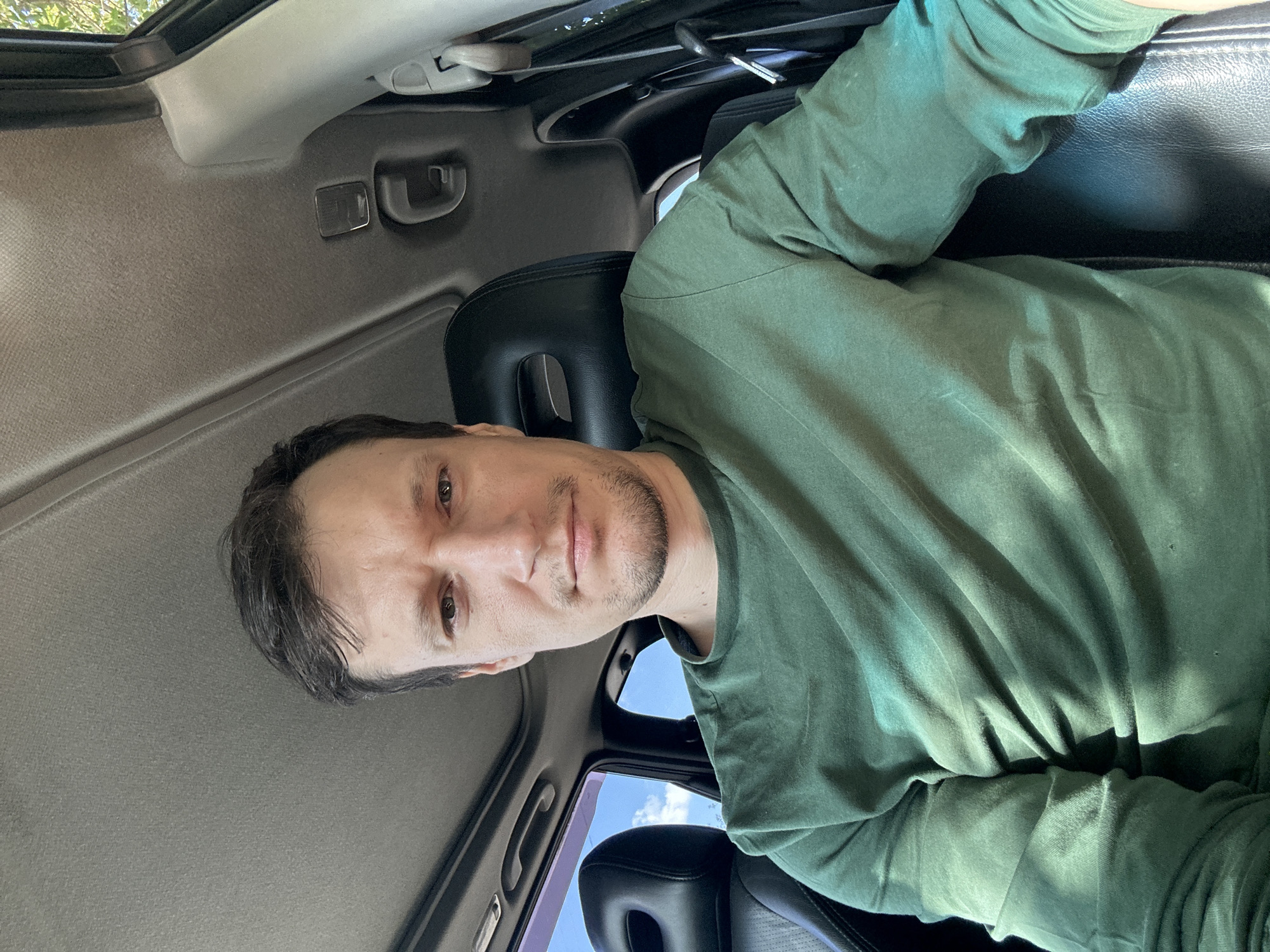 A man with short, dark hair and stubble is pictured from the chest up, sitting in the driver's seat of a car. He is wearing a long-sleeved green t-shirt. His expression is neutral. The car interior, with black leather seats and a sunroof, is visible. The exterior environment, seen through the car window, suggests a sunny day with blue sky and trees. The location is identified as Makiv, Ukraine.