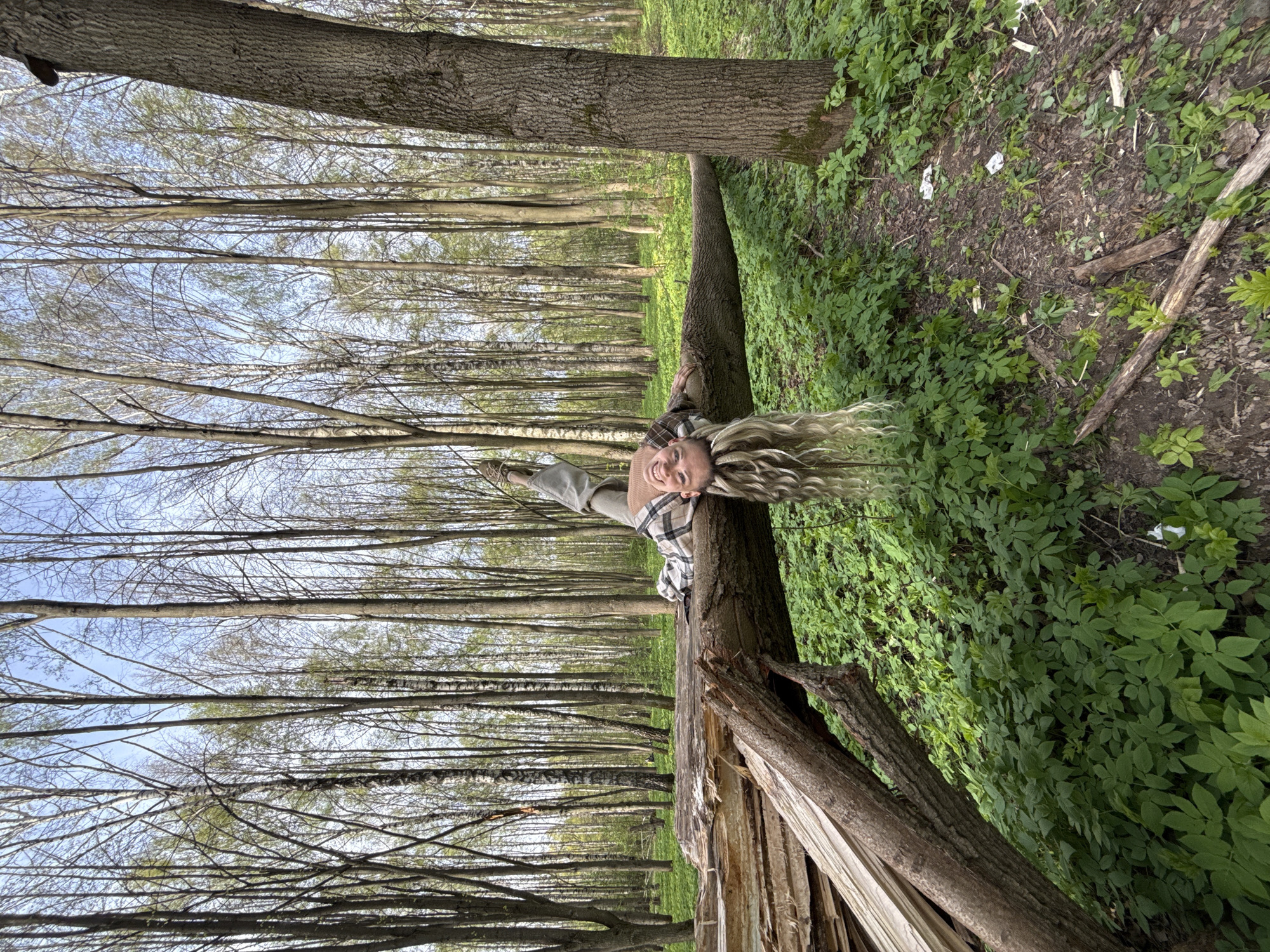 The image depicts a woman lying across a fallen tree in a forest. The tree is split near its base, exposing light-colored wood. The surrounding environment consists of tall, thin trees with bare branches, a ground cover of green plants, and leaf litter. The woman has blonde hair and is wearing a plaid shirt and grey sweater. The location is identified as Rusanovo, Russia.