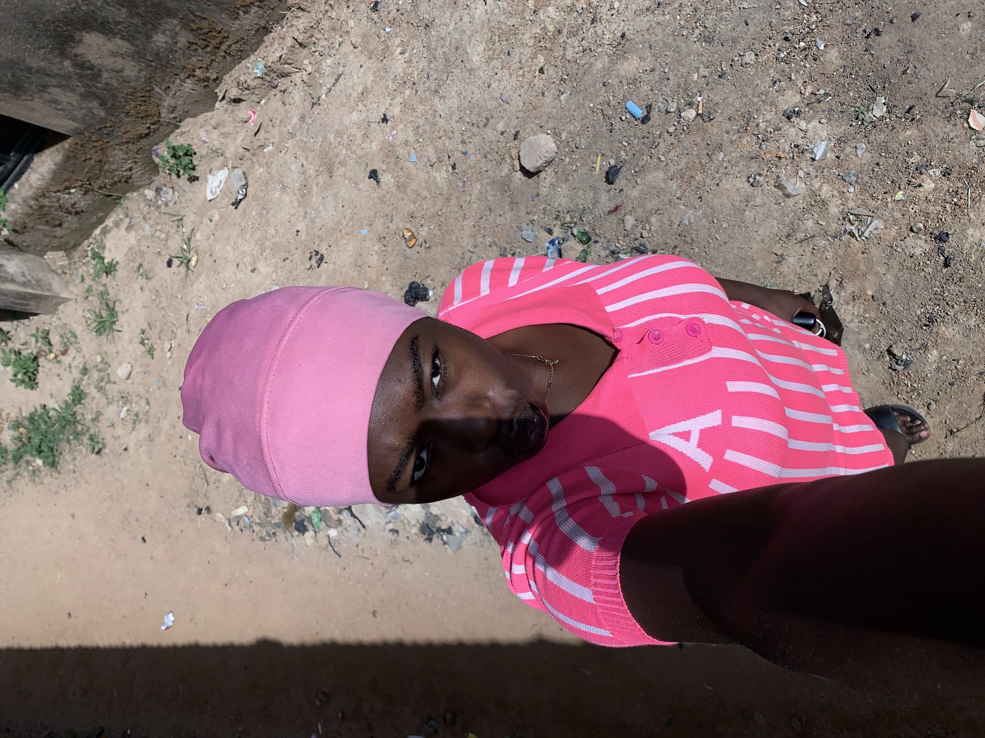 A person wearing a pink head covering and a pink and white striped shirt is photographed from a high angle in Rafin Par, Nigeria. The ground is dirt and littered with small debris. A building wall is visible on the left side of the frame. The person appears to be looking up at the camera.