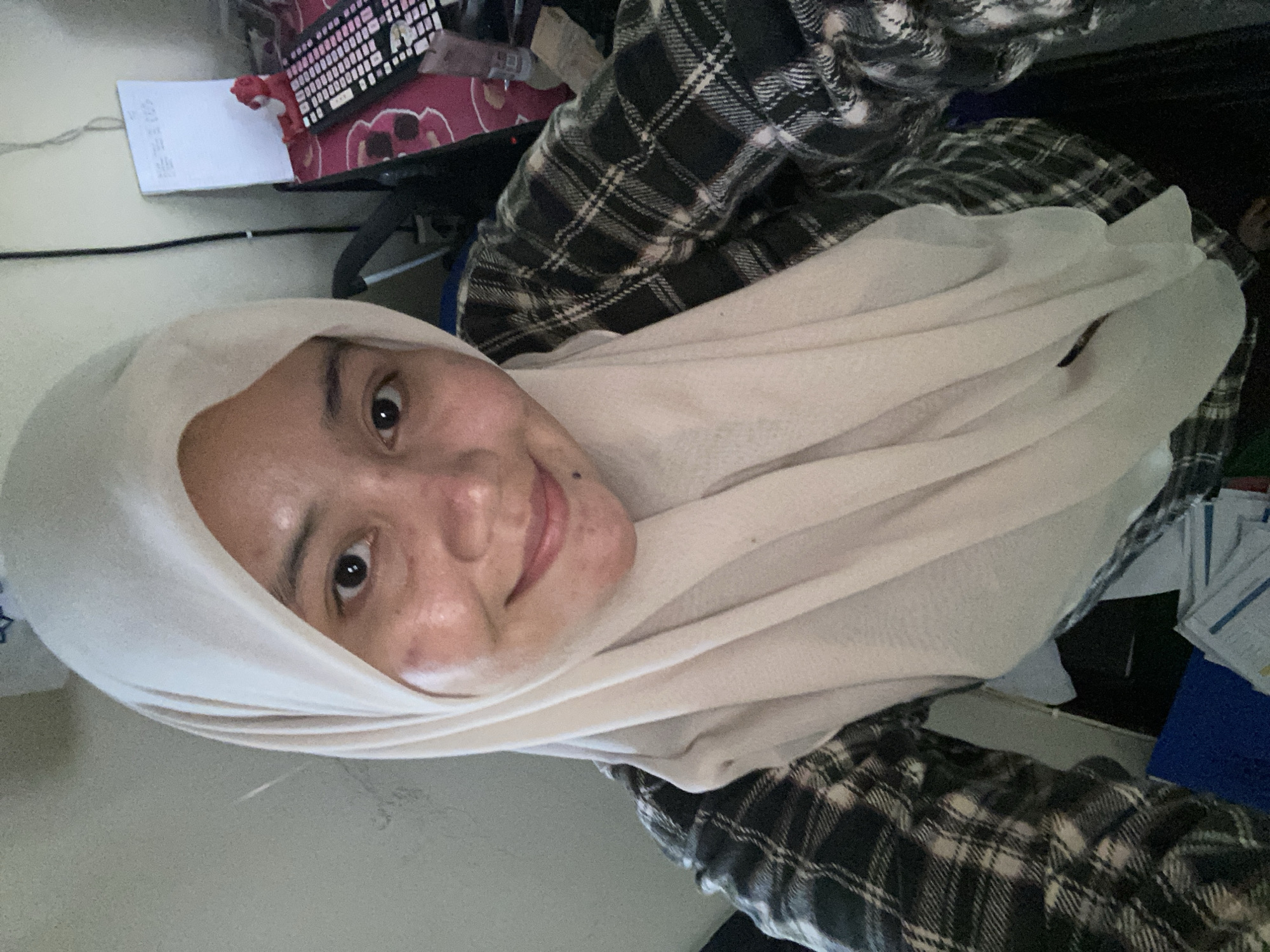 The image shows a close-up, selfie-style shot of a young woman wearing a light-colored hijab and a dark plaid shirt. She is looking directly at the camera and smiling. Her skin appears to have some blemishes and is somewhat shiny. In the background, there is a white board with some writing on it, a red figurine, and a keyboard. The overall setting appears to be an indoor environment, possibly a room or office. The image does not provide any specific visual cues to definitively place it in Banjarbaru, Indonesia, or any other specific city.