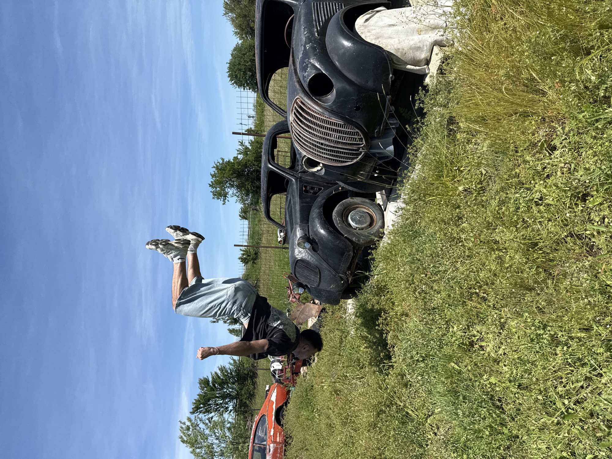 A man is performing a backflip in a grassy field in Omurtag, Bulgaria. The man is wearing shorts and a black t-shirt. Behind him are two old, rusted cars and a metal fence. The sky is clear and blue.