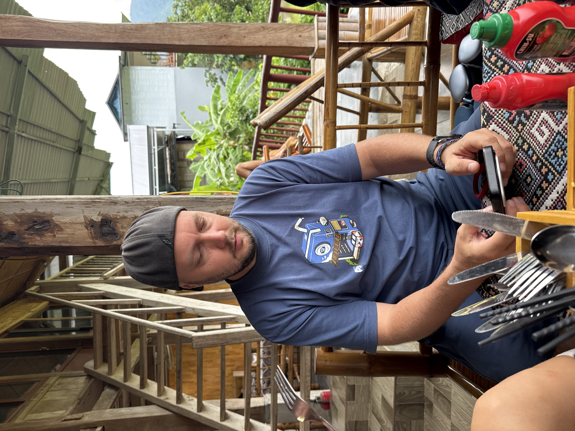 In Xóm Khán, Vietnam, a man wearing a grey baseball cap and a blue t-shirt with a cartoon graphic sits at a table on a wooden porch. He is holding a black object in his hands and looking down at it. A variety of cutlery, including knives and forks, are arranged in a wooden holder on the table in front of him. Two bottles, one red and one green, are also visible on the table. In the background, a wooden staircase leads up to a building, and lush green foliage is visible. The scene suggests a casual outdoor dining setting in a rural or semi-rural area.