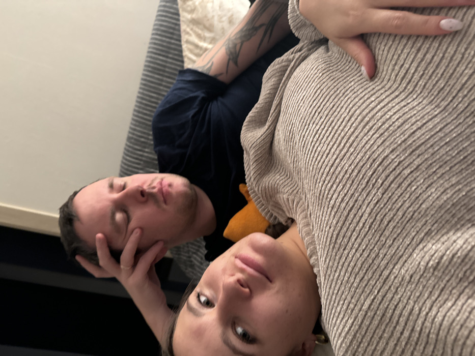 This image captures two individuals reclining indoors, likely on a sofa or bed, in Селекционер-2 Снт, Russia. In the foreground, a female subject with light brown hair and a fair complexion is seen lying on her back, angled towards the camera. She wears a beige ribbed long-sleeved top and a gold-colored hoop earring in her visible left ear. Her left hand, with light-colored fingernail polish featuring small dark specks, rests on her mid-section. Her eyes are open, directed towards the viewer, and her mouth is closed.
Behind her and to the right, a male subject with short dark hair and a fair complexion is also reclining. His eyes are closed or heavily lidded, and his right hand is raised, with fingers resting against his right temple. A simple band-style ring is visible on his left ring finger. He wears a dark blue short-sleeved t-shirt, revealing a sleeve tattoo on his left arm that displays dark, intricate patterns, possibly depicting birds or foliage. An orange piece of fabric, possibly a cushion, is positioned between the two subjects. The background shows a striped grey and white upholstered surface, likely a sofa back, and a light-colored wall. A darker vertical element is visible on the far left of the frame.