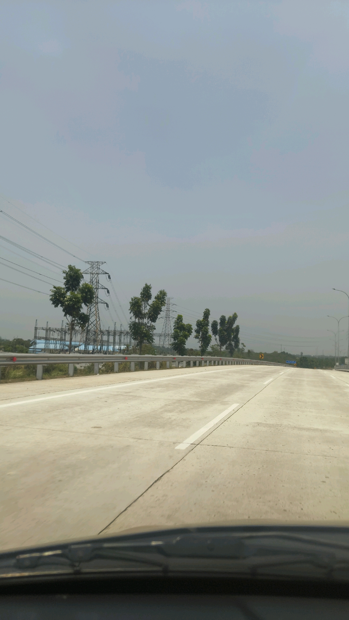 The image depicts a highway in Situ Gadung, Indonesia, under a hazy sky. The perspective is from the interior of a vehicle, showing the windshield and dashboard in the foreground. The highway itself is made of concrete with white lane markings. On the left side of the highway, there are several tall electrical transmission towers with wires stretching across the landscape. Between the towers and the highway, a metal guardrail is visible, with sparse vegetation behind it. A line of trees with rounded canopies stands in front of the transmission towers. In the distance, more trees and low-lying structures can be seen. There are no people visible in the image.