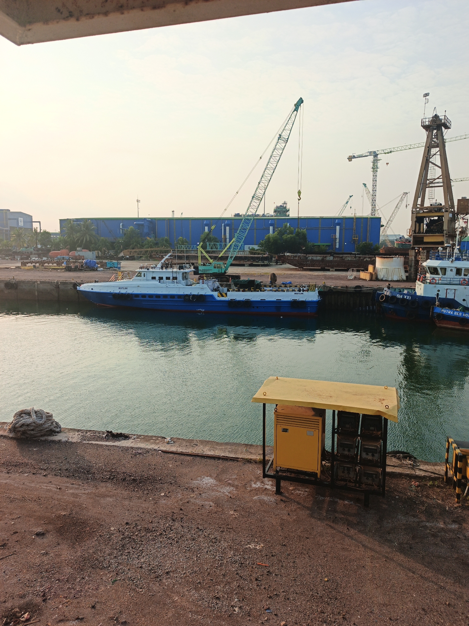 The image depicts a port or shipyard area in Batam, Indonesia. In the foreground, a concrete dock is visible with a large coiled rope on the left and a brown, gravelly ground surface. A yellow metal shelter with electrical equipment is situated on the dock. The middle ground shows a body of water with several vessels docked. A prominent white and blue boat, labeled "RESTLINK," is moored to the left. To the right, two other boats are docked, one dark blue with "SM XXI" on its side and another partially visible labeled "PUTRA BES DDA." In the background, a large blue industrial building stands, with several construction cranes of various sizes and colors scattered around the area. The sky is overcast, suggesting either early morning or late afternoon.