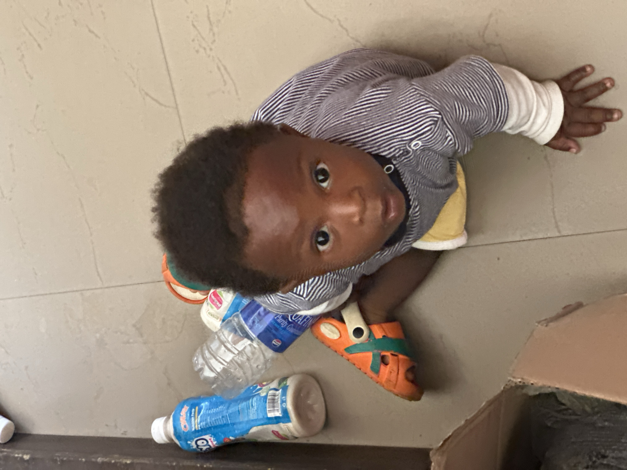 In this overhead shot, a young child with dark hair and eyes looks up at the camera from a seated position on a tiled floor. The child is wearing a horizontally striped long-sleeved shirt and orange sandals with green accents. Their left hand is raised, fingers splayed, resting on the floor. Several plastic bottles are scattered around the child, some appearing to be water bottles and one a blue drink bottle with visible text that is partially obscured but includes "O" and "250ml". A cardboard box is visible in the lower right corner of the frame. The lighting suggests it is daytime. The setting appears to be indoors, possibly a home or public space with simple tiled flooring. The child's gaze and raised hand could indicate curiosity or a playful interaction. There are no obvious indicators of the weather or specific time of day beyond daylight.