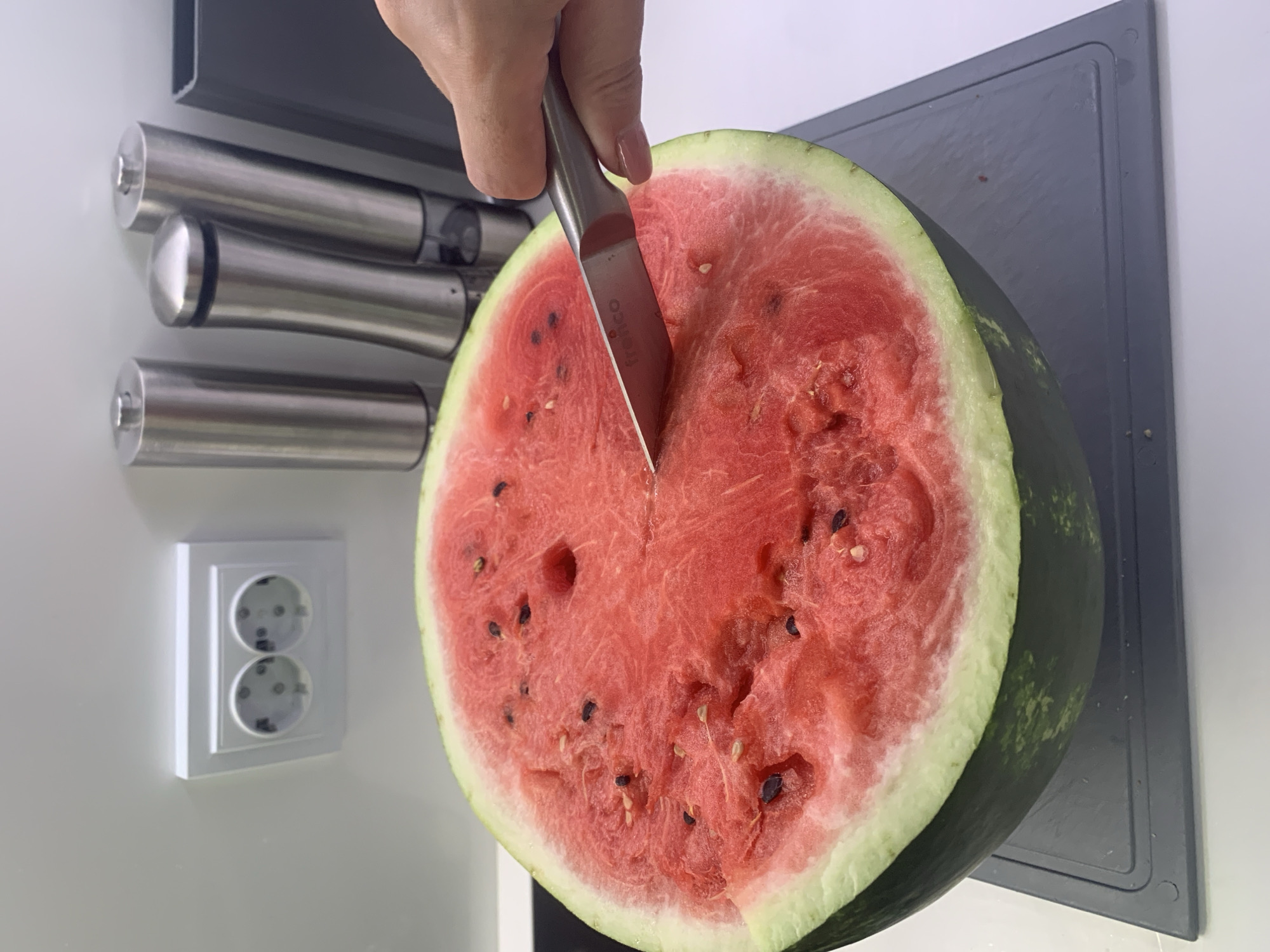 A half watermelon is being cut by a knife held by a person's hand in what appears to be a kitchen. The watermelon is red with black seeds and a green rind, resting on a grey cutting board. In the background, stainless steel salt and pepper grinders are visible, along with a white electrical outlet. The overall scene is a close-up, indoor shot.