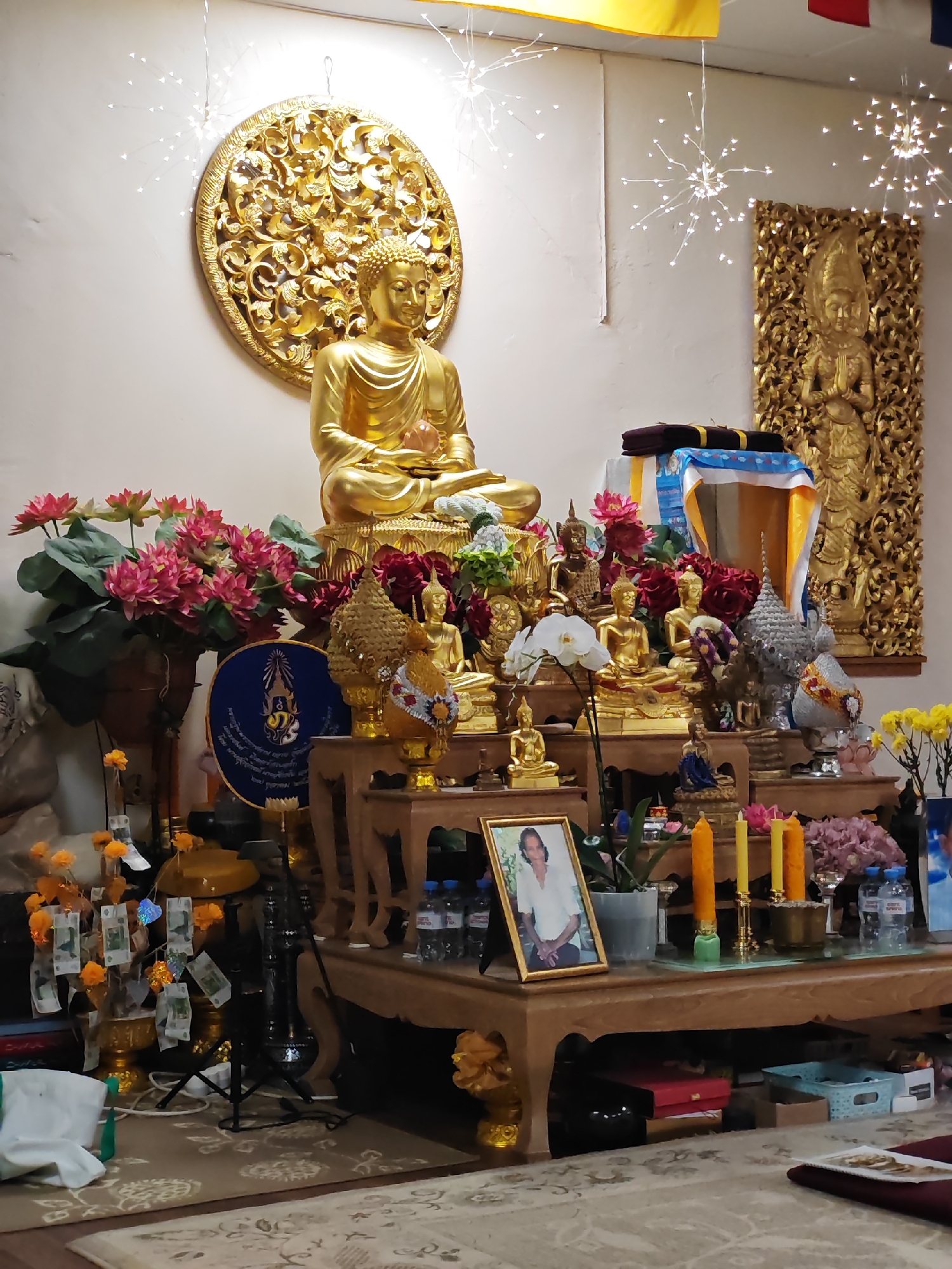 The image shows a Buddhist shrine in an indoor setting. The shrine consists of several golden Buddha statues of varying sizes placed on tiered wooden platforms. The largest Buddha statue sits atop the highest platform, framed by a circular golden floral carving on the wall behind it. A photograph in a golden frame is visible on the lower platform. Other objects on the shrine include flowers, candles, water bottles, and offerings. The setting appears to be a temple or place of worship with ornate golden carvings adorning the walls. The presence of currency clipped to small trees suggests devotional offerings. There are light fixtures resembling starbursts hanging from the ceiling, providing illumination. The image does not contain verifiable information regarding the city of Saint Petersburg, Russia.