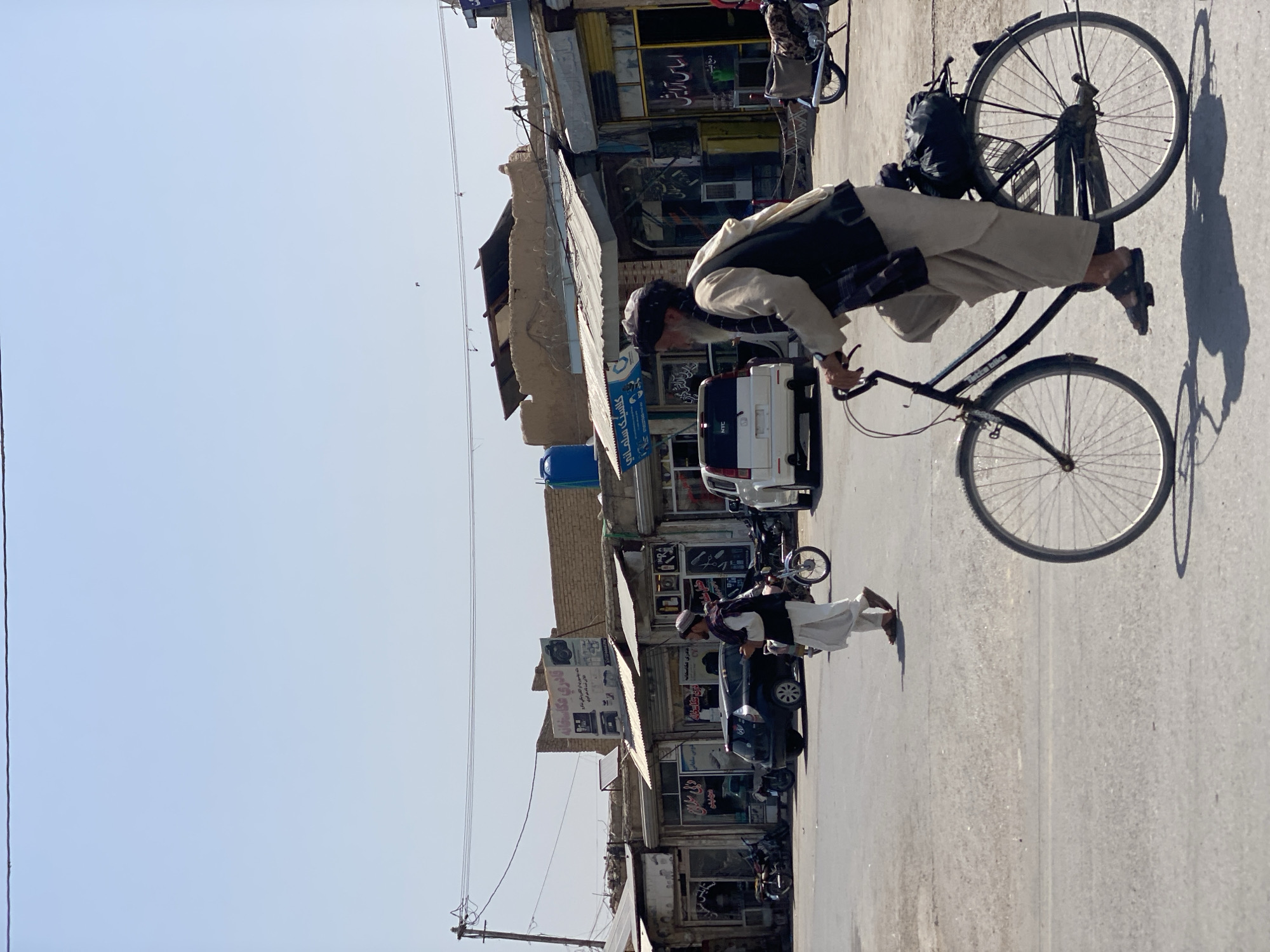The image captures a street scene in Kandahar, Afghanistan, on a bright, clear day. In the foreground, an elderly man rides a bicycle. He is wearing traditional Afghan clothing, including a loose, light-colored tunic and trousers, a vest, and a dark turban. A black bag is secured to the back of his bicycle. The man's face is weathered, and he appears to be looking forward as he pedals.
Behind him, the street is paved and appears to be a commercial area. Several buildings with awnings line the street, housing various shops. Signs in Pashto and possibly other local languages are visible. One sign features an image of a camera and text that appears to indicate a photography studio. Another sign has Arabic script, possibly indicating a beauty salon. In the middle ground, other people are present. One man, also dressed in traditional clothing, walks away from the camera, while another man is visible walking near a parked car. Several motorcycles and cars are parked along the street, including a black SUV and a white van. The architecture of the buildings suggests a blend of older and newer construction. The overall impression is of a bustling urban environment in Afghanistan.