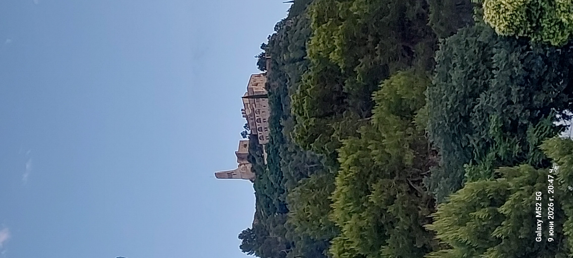 The image shows the Montserrat monastery complex, located on the mountain of the same name, in the municipality of Monistrol de Montserrat, within the province of Barcelona, Catalonia, Spain. The monastery, perched atop a lush, tree-covered hillside, is partially visible under a clear blue sky. A prominent, slender tower stands out on the left side of the complex, while other stone buildings with arched windows and tiled roofs are visible to its right. The foreground is dominated by dense green foliage of various trees and shrubs, obscuring the base of the hill. A timestamp in the bottom left corner reads "Galaxy M52 5G 9 юни 2026 г. 20:47 ч.", indicating the photograph was taken on June 9, 2026, at 8:47 PM.