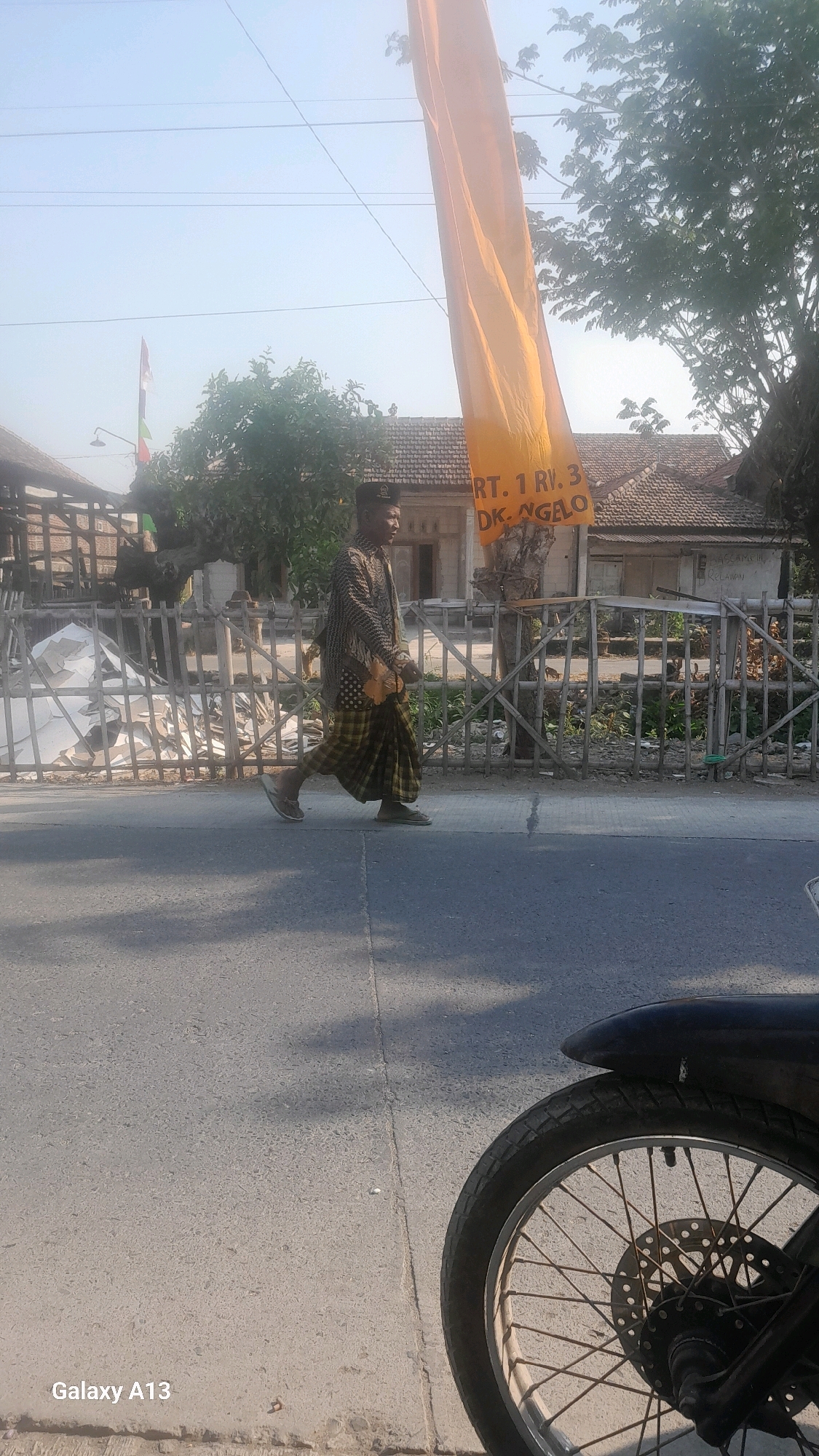 In Ngelo, Indonesia, under a clear, bright sky, a man walks along a paved road. He is wearing a traditional patterned shirt, a checkered sarong, a black cap, and flip-flops. He carries a small bag on his left shoulder. Behind him, a wooden fence lines the road, with debris and building materials visible on the other side. A large orange banner hangs from a tree, displaying text that includes "RT. 1 RV. 3 DK. NGELO." In the foreground, the wheel and fender of a motorcycle are visible, suggesting the photo was taken from a stationary motorcycle. The scene depicts a typical day in a residential area of Indonesia.