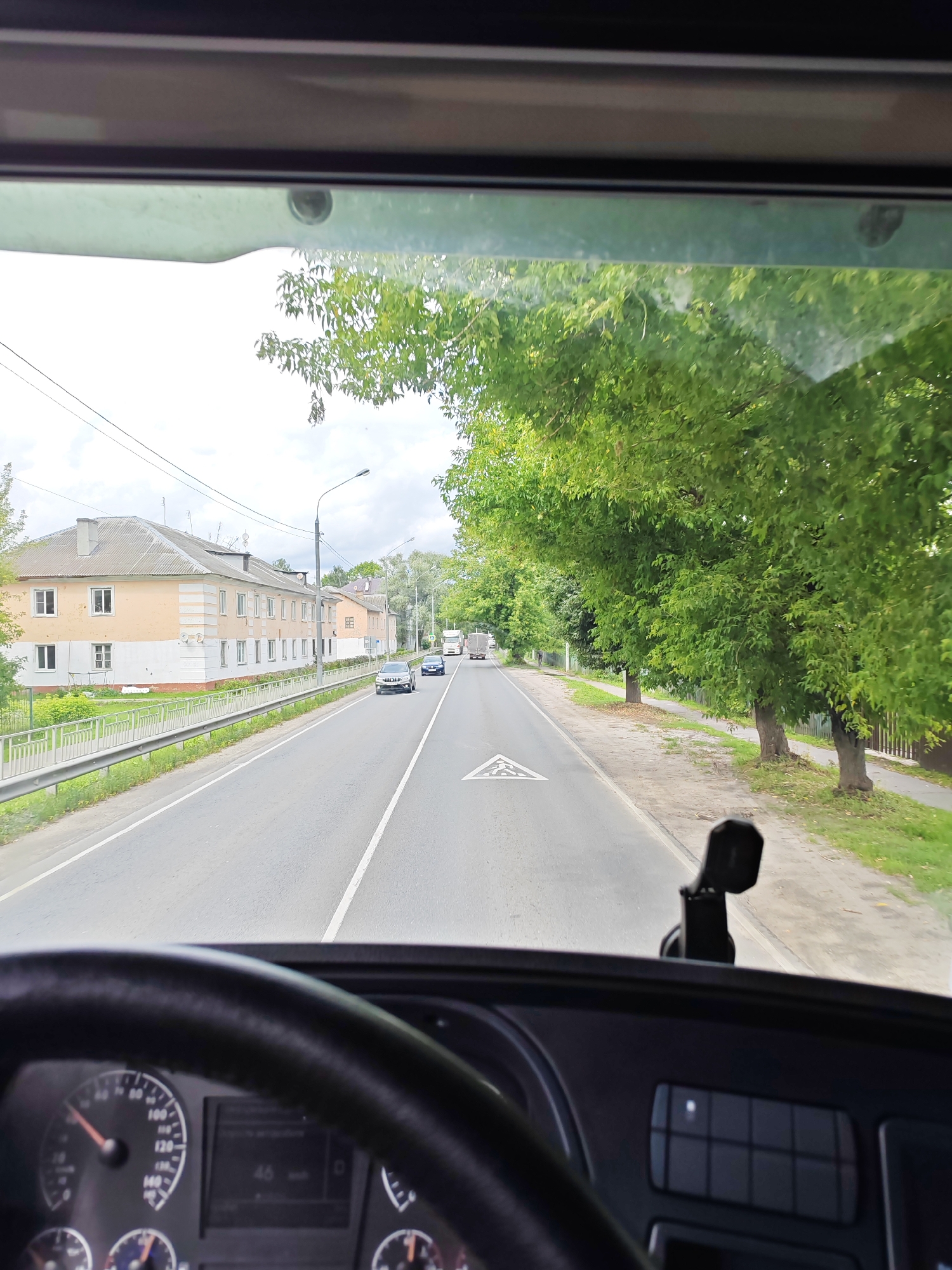 The image captures a view from the driver's perspective inside a vehicle, likely a truck or bus, as it travels along a two-lane road in Likino-Dulyovo, Russia. The road appears to be asphalt with clear white lane markings. On the left side of the road, a row of multi-story residential buildings with light yellow and white facades and many windows is visible. A metal guardrail separates the road from a grassy area in front of the buildings, which is further bordered by a low fence. Across the road, a sidewalk runs alongside a dense line of mature green trees. Several vehicles are present on the road, including passenger cars and larger trucks in the distance. The sky is overcast with some visible clouds. In the foreground, the steering wheel and dashboard of the vehicle are prominent, with a speedometer showing approximately 46 km/h.